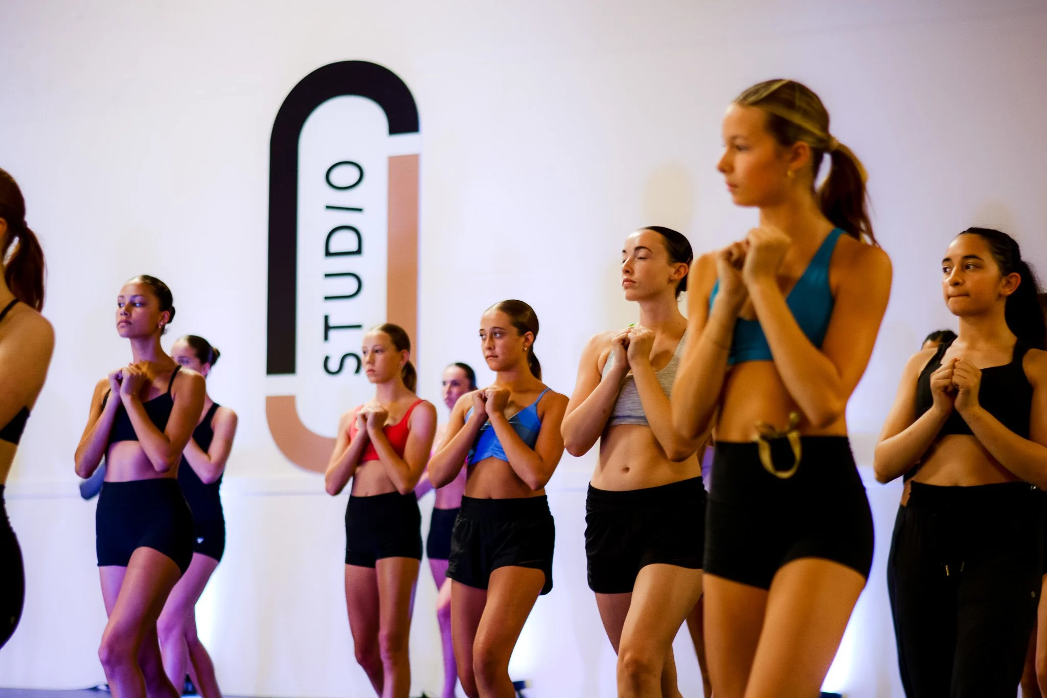 Line of young women in workout clothes standing with hands clasped to their chests during a dance class or rehearsal at Studio J.