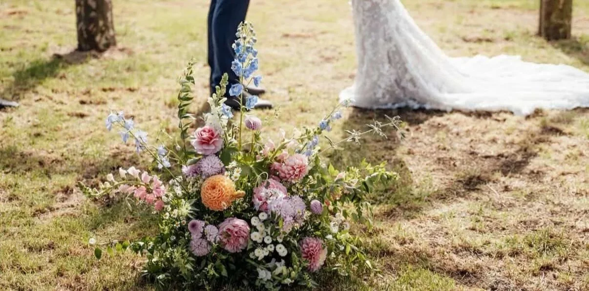 A floral arrangement with pink and white flowers in a white textured vase, next to a jar with string lights, on a table with a green table runner, in a softly lit setting with bokeh lights in the background.
