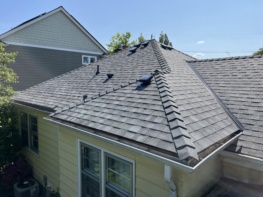 Close-up of a residential house roof with gray asphalt shingles, skylights, vents, and a gutter system, under a bright blue sky with a neighboring house visible in the background.