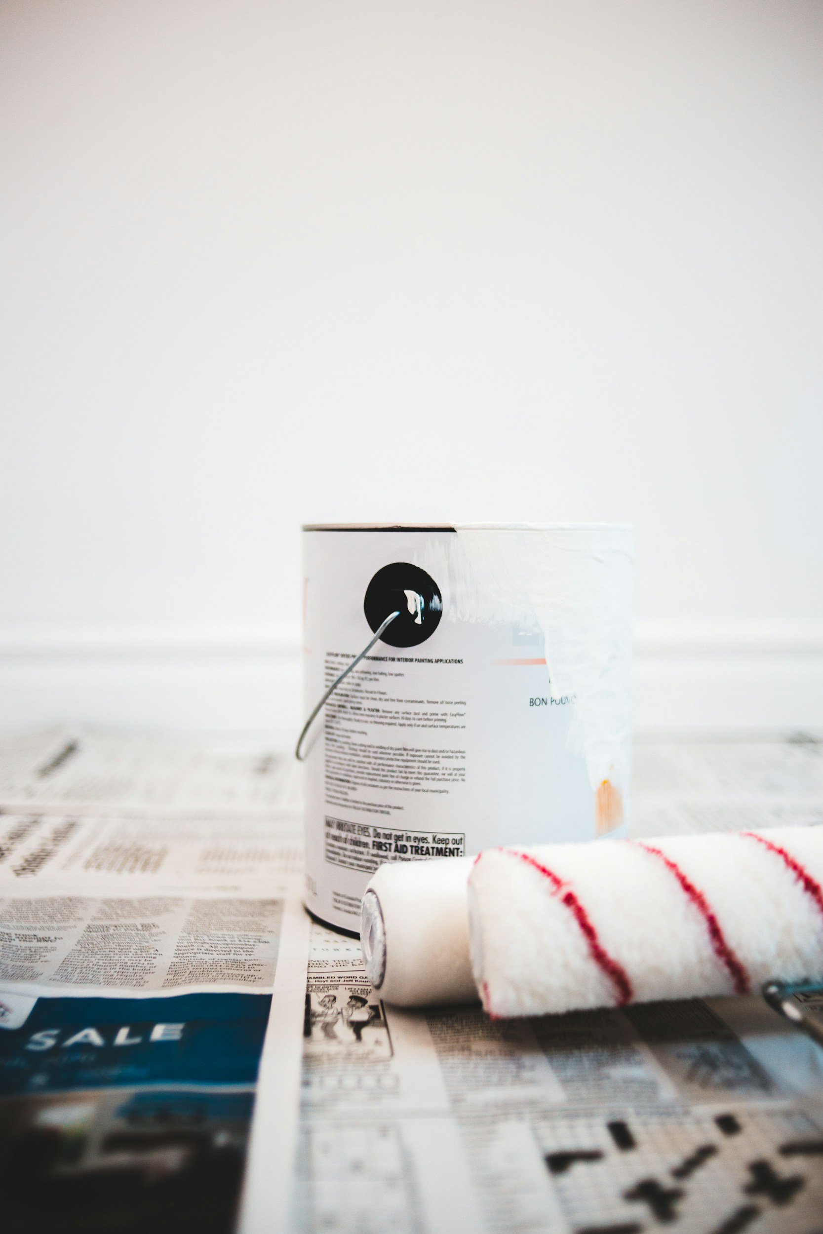 A container of interior paint, a paint roller with a red and white striped cover, and a rolled-up piece of masking tape on newspapers, all set against a plain white background.