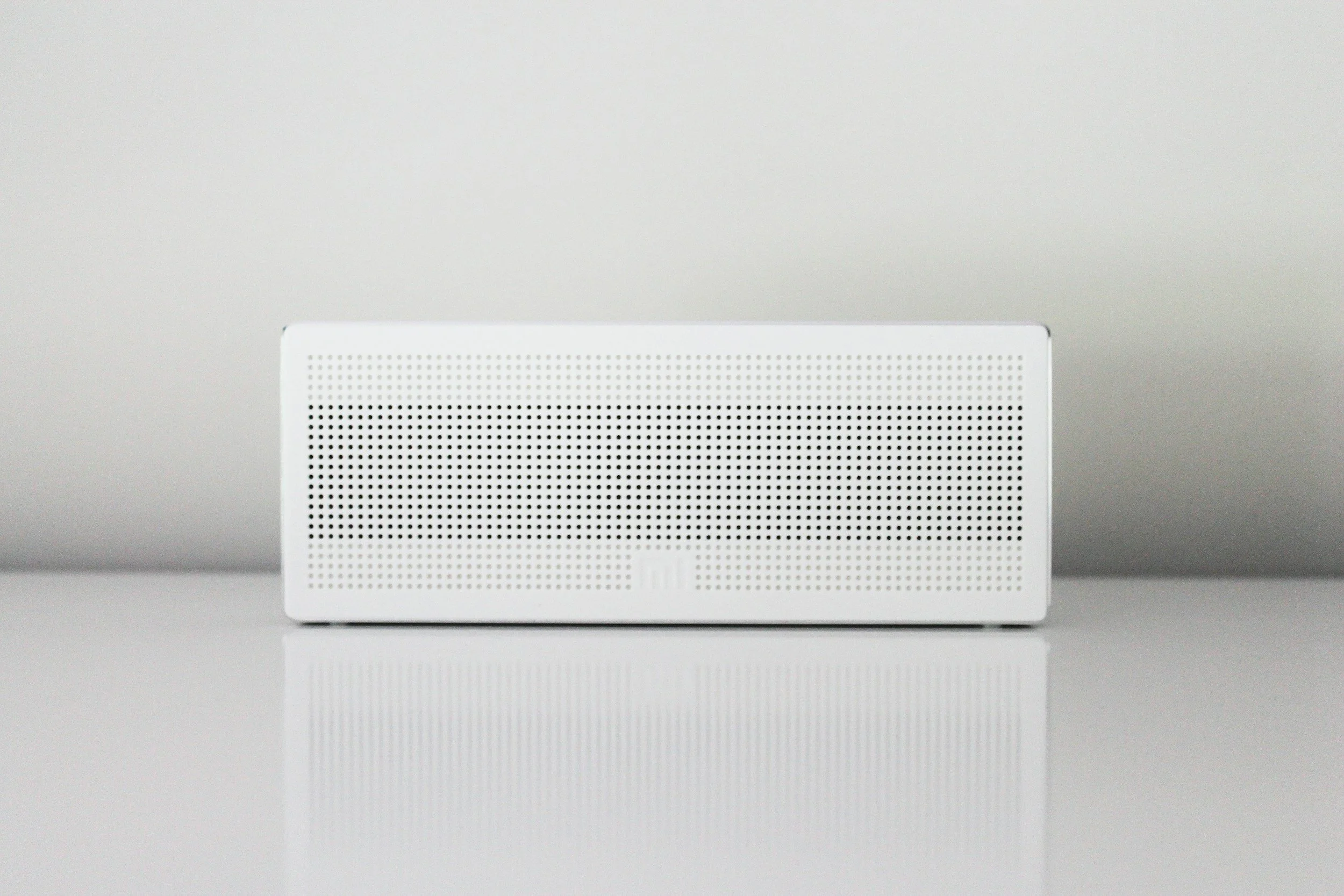 White rectangular smart speaker with perforated front grille on a reflective surface against a plain light gray background.