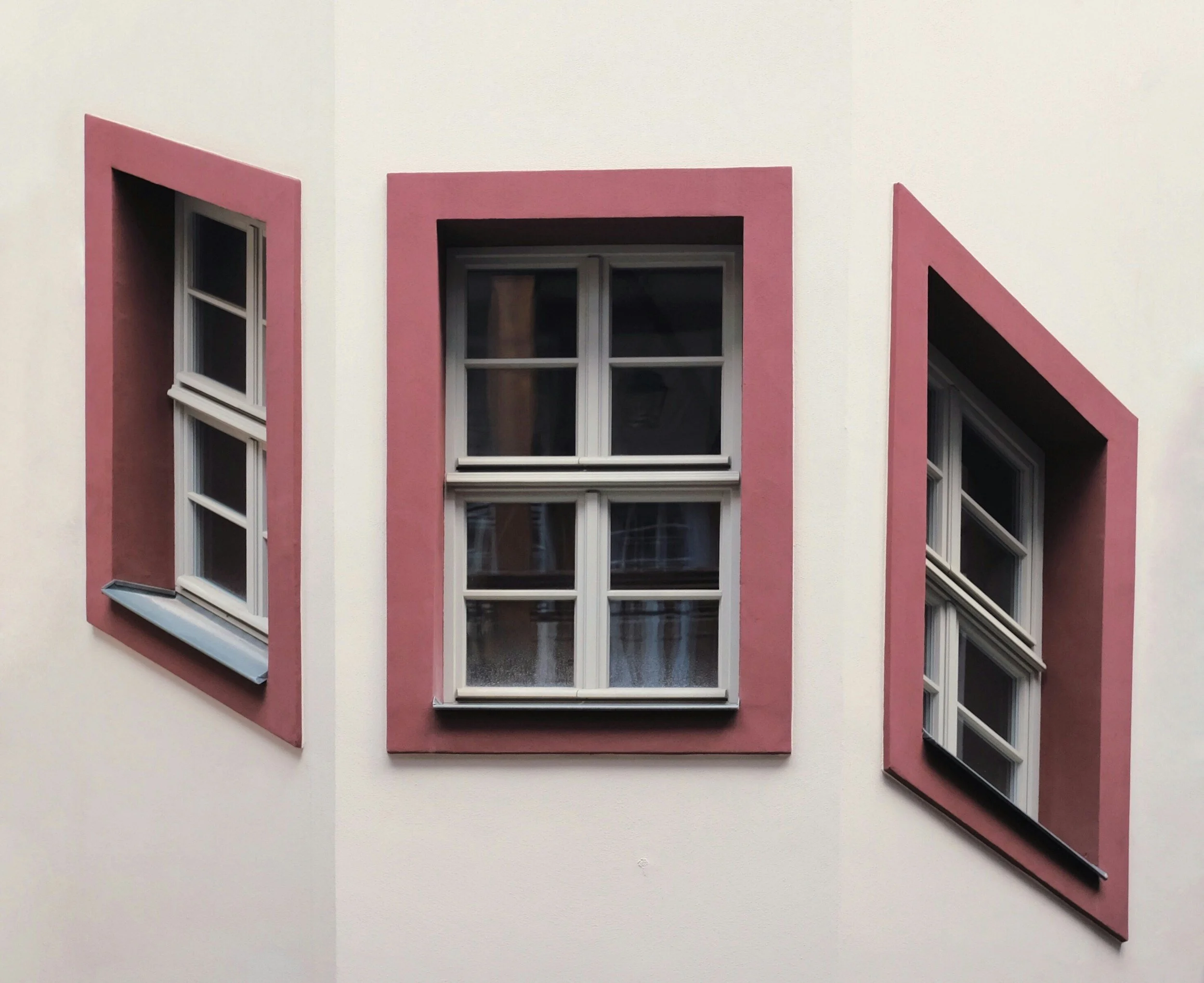 Three windows with white frames on a white building, each with pink trim around them. The windows are different sizes and styles, with the middle one being the largest. The right window has its shutters partially open, and the pink trim around each window is slanted, creating a visual pattern.