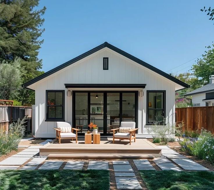 Modern white house with a black roof, outdoor seating on a wooden deck, surrounded by a landscaped yard with plants and stone pathway.
