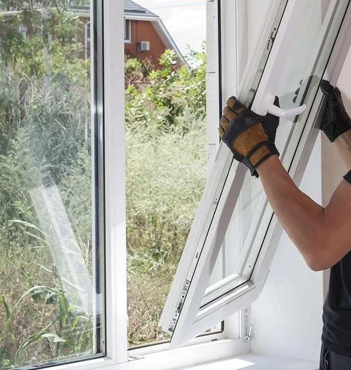 A person wearing work gloves installing a white window frame inside a house.