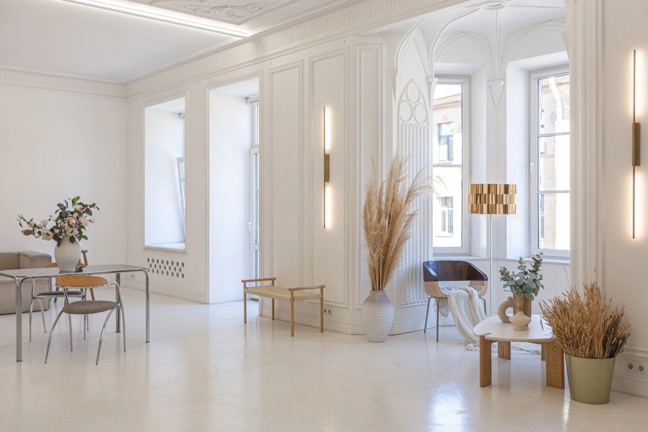 A bright, minimalist interior room with white walls, large windows, and simple decor including a vase with flowers, a small table, and modern lighting fixtures.