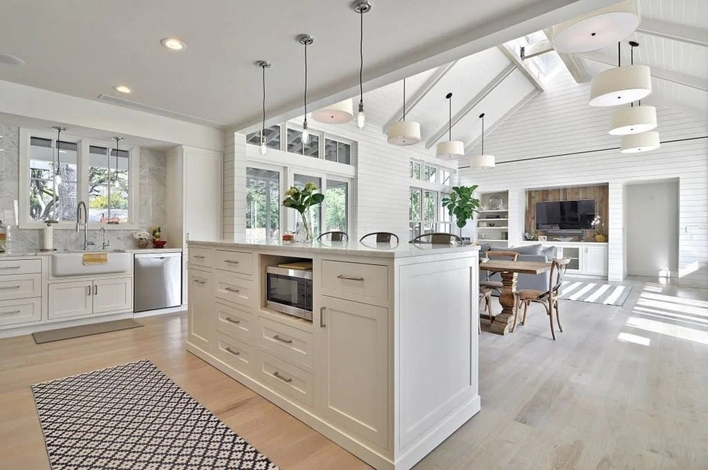 Bright open-concept kitchen and living area with white cabinetry, large windows, wooden dining table, TV, and pendant lights.