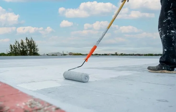 A person is using a paint roller to apply gray waterproof coating on a flat roof outdoors, with a cloudy sky and trees in the background.