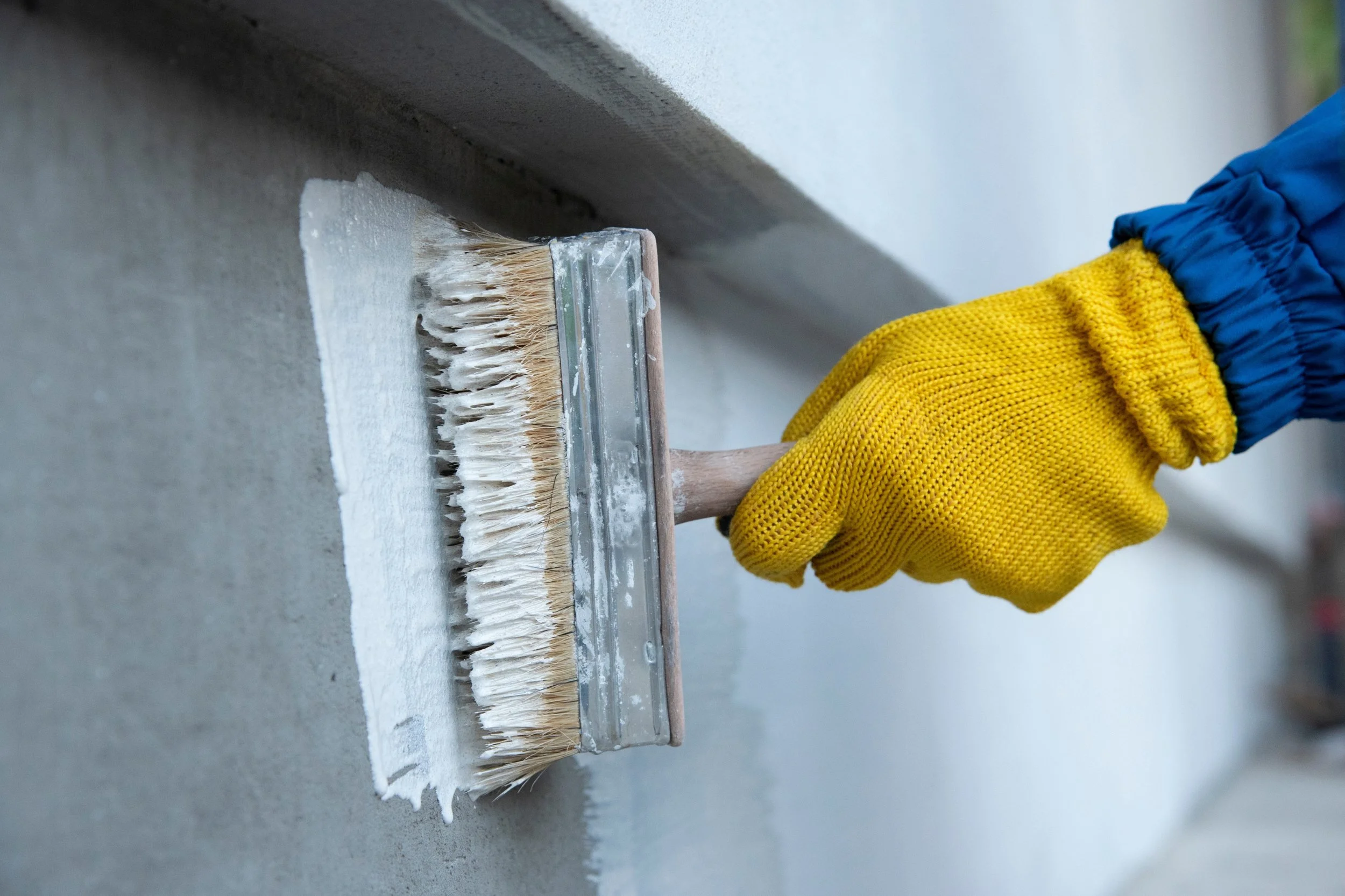 Person wearing yellow gloves and a blue sleeve holds a wide brush applying white paint to a wall.