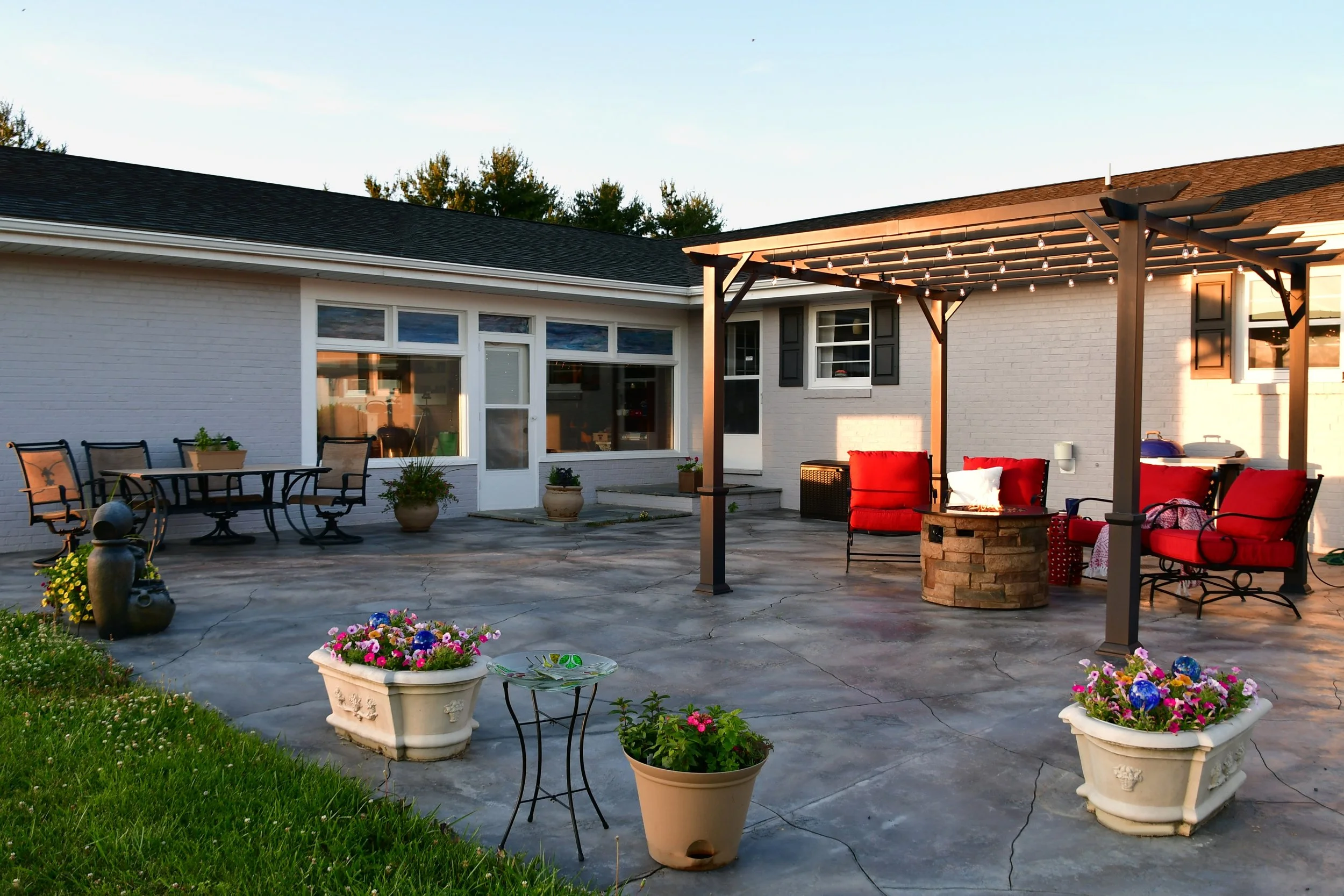 Backyard patio with outdoor furniture, potted plants, and string lights, attached to a house with white brick walls and large windows, during sunset.