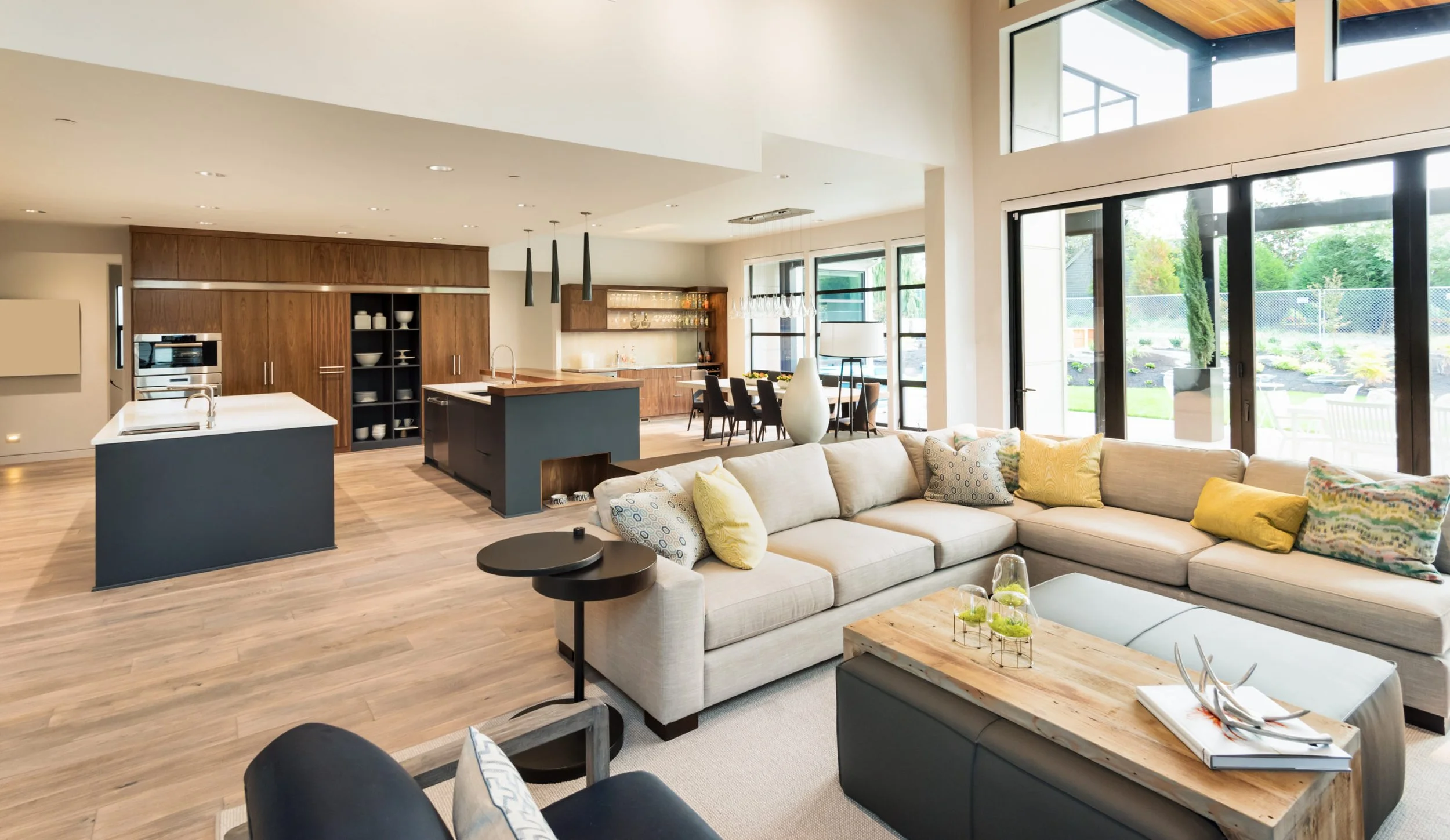 Open-concept living room and kitchen with large windows, beige sectional sofa, wooden coffee table, and modern kitchen with island and dining area.