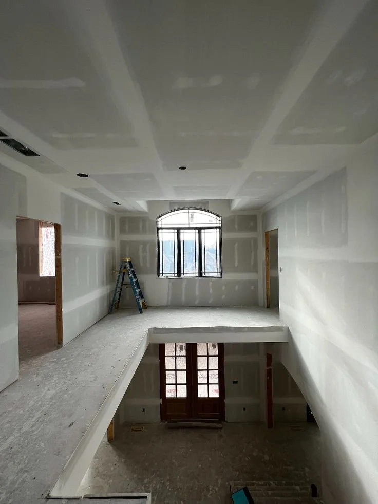 Interior of a house under construction with drywall installed, a ladder, a large arched window, and a door with glass panels leading outside.