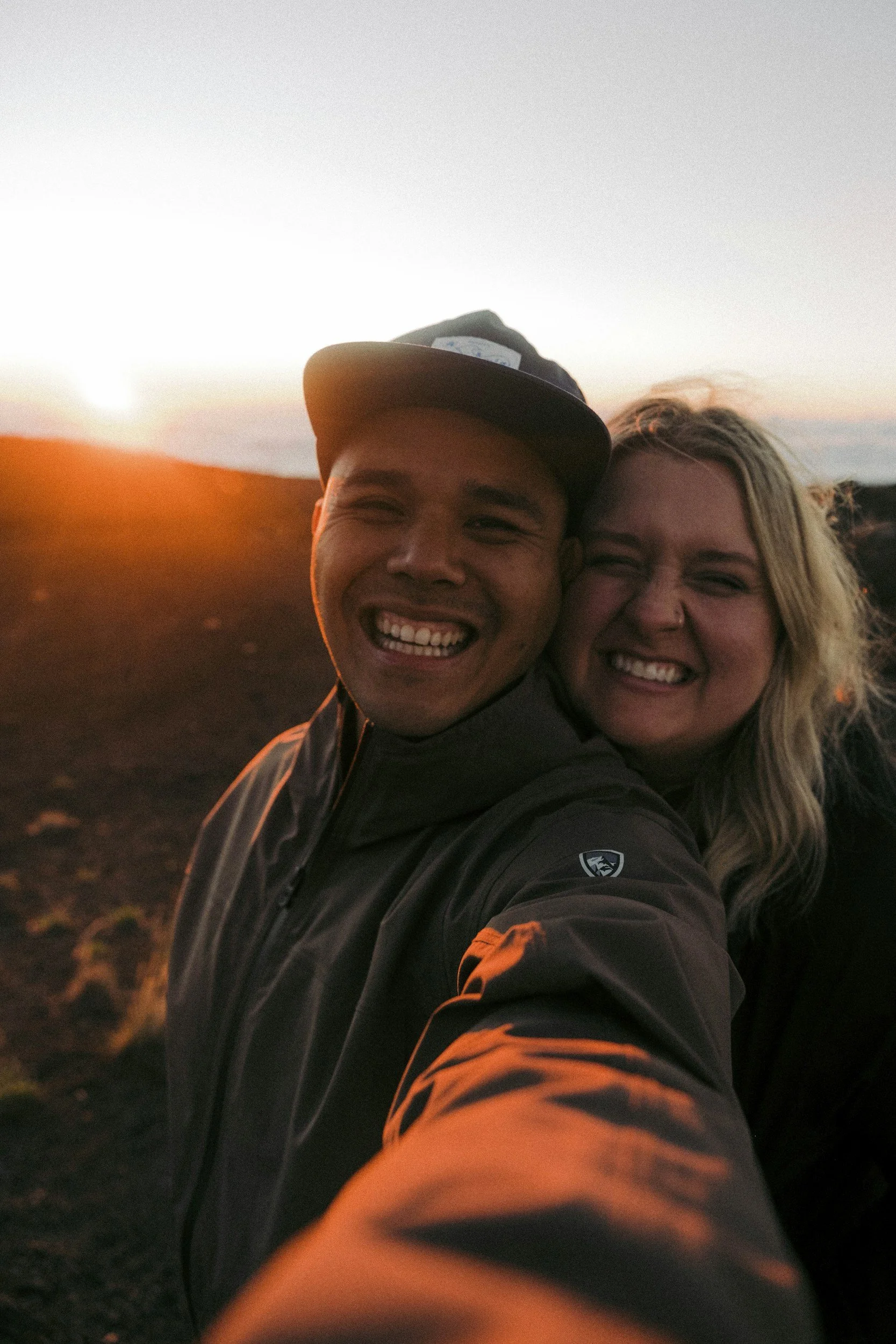 A couple taking a selfie at sunset outdoors, smiling and close together with a landscape in the background.