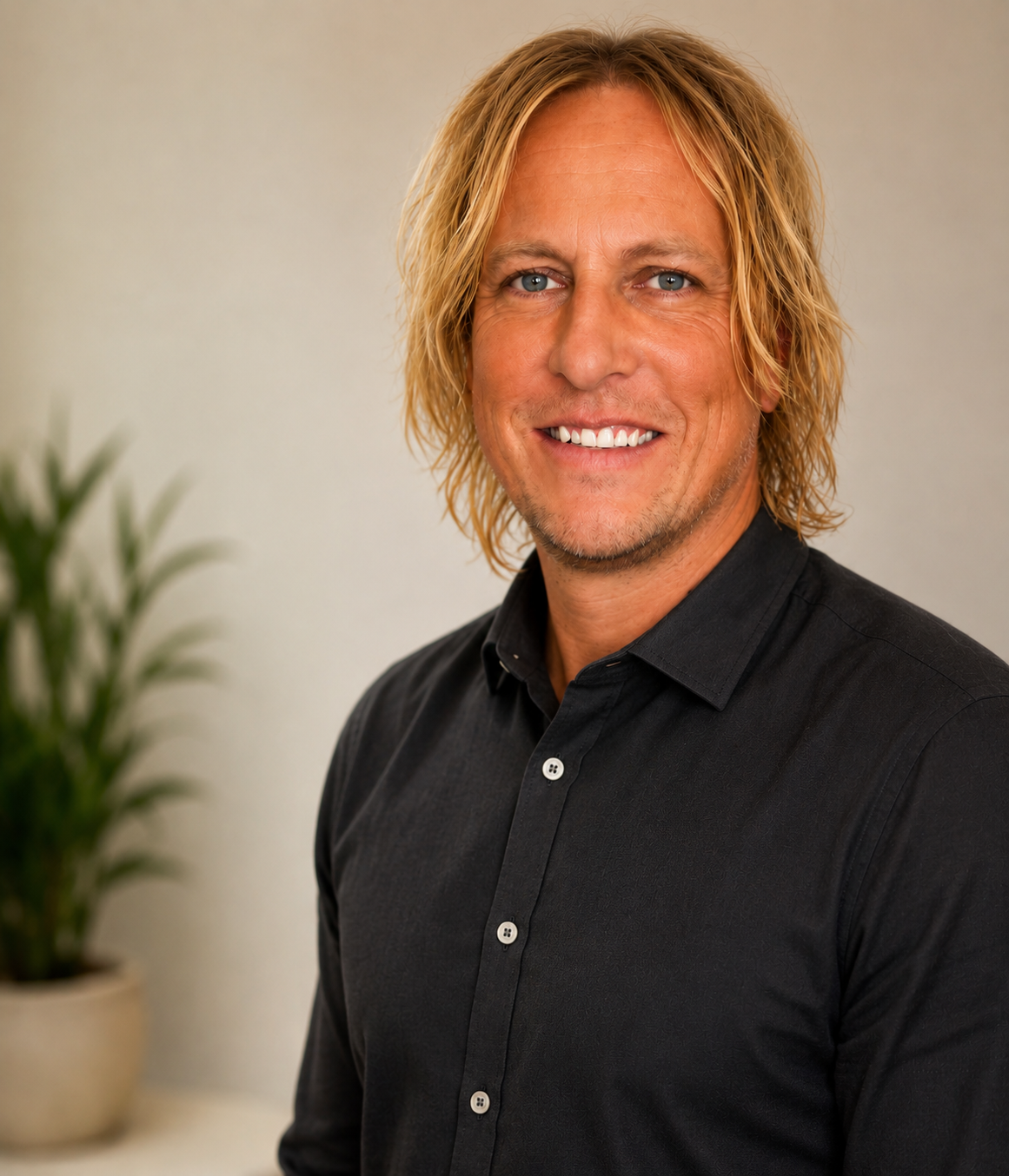 Headshot of a smiling man with shoulder-length blond hair wearing a black button-up shirt, standing in front of a light-colored wall and a potted plant.