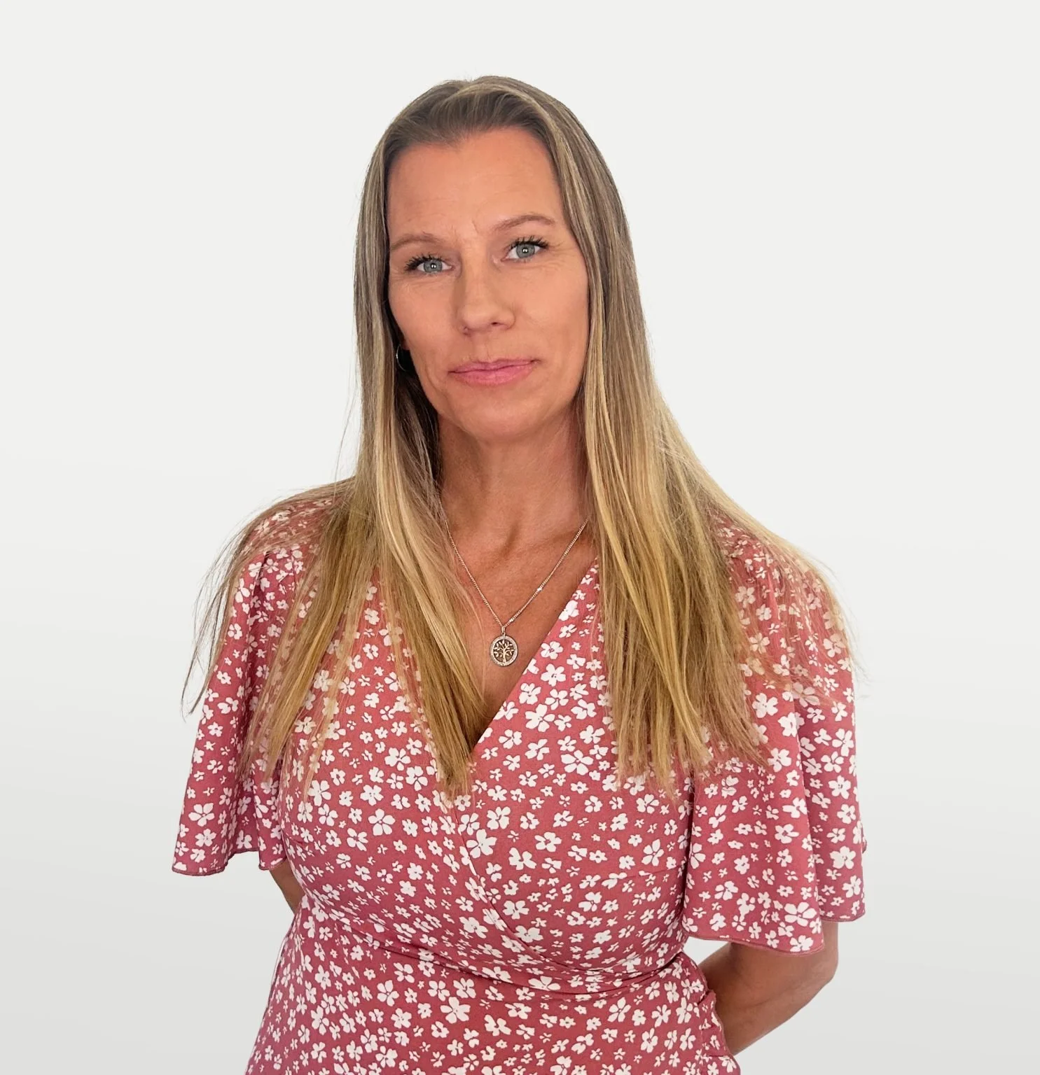 A woman with long blonde hair and blue eyes wearing a pink dress with white floral patterns and a silver necklace with a tree pendant, standing against a plain white background.