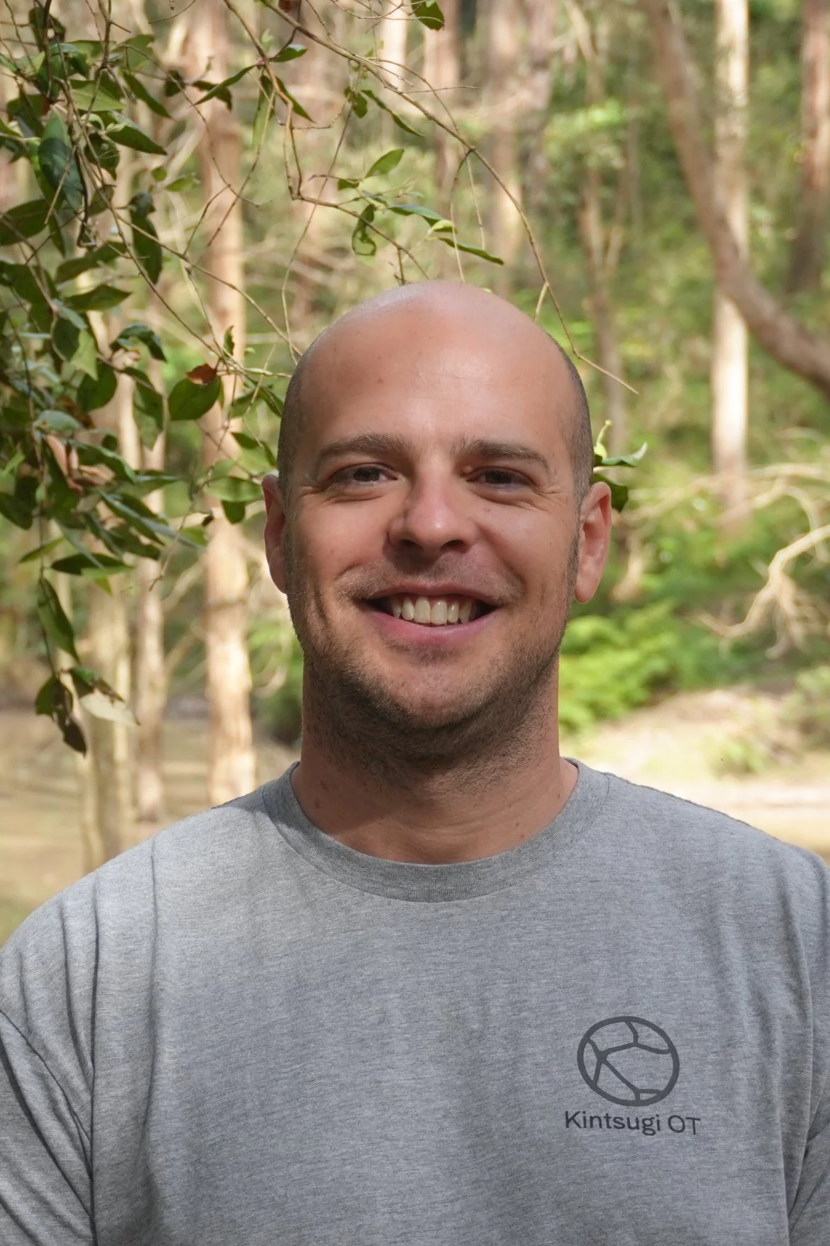 A smiling bald man in a gray T-shirt standing outdoors in a wooded area.