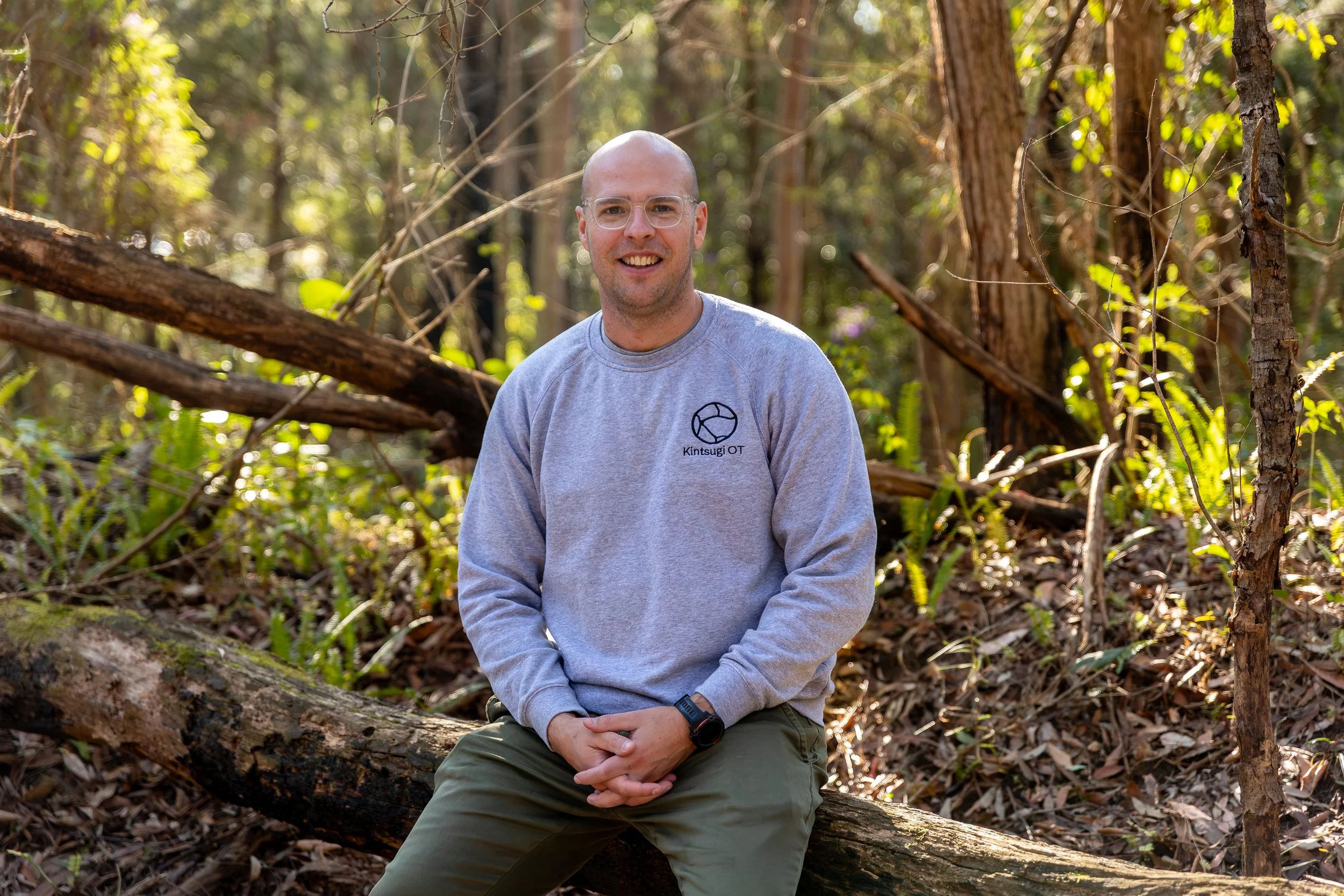 A man with glasses and a gray sweatshirt sitting on a fallen tree in a wooded area with trees and green foliage in the background.