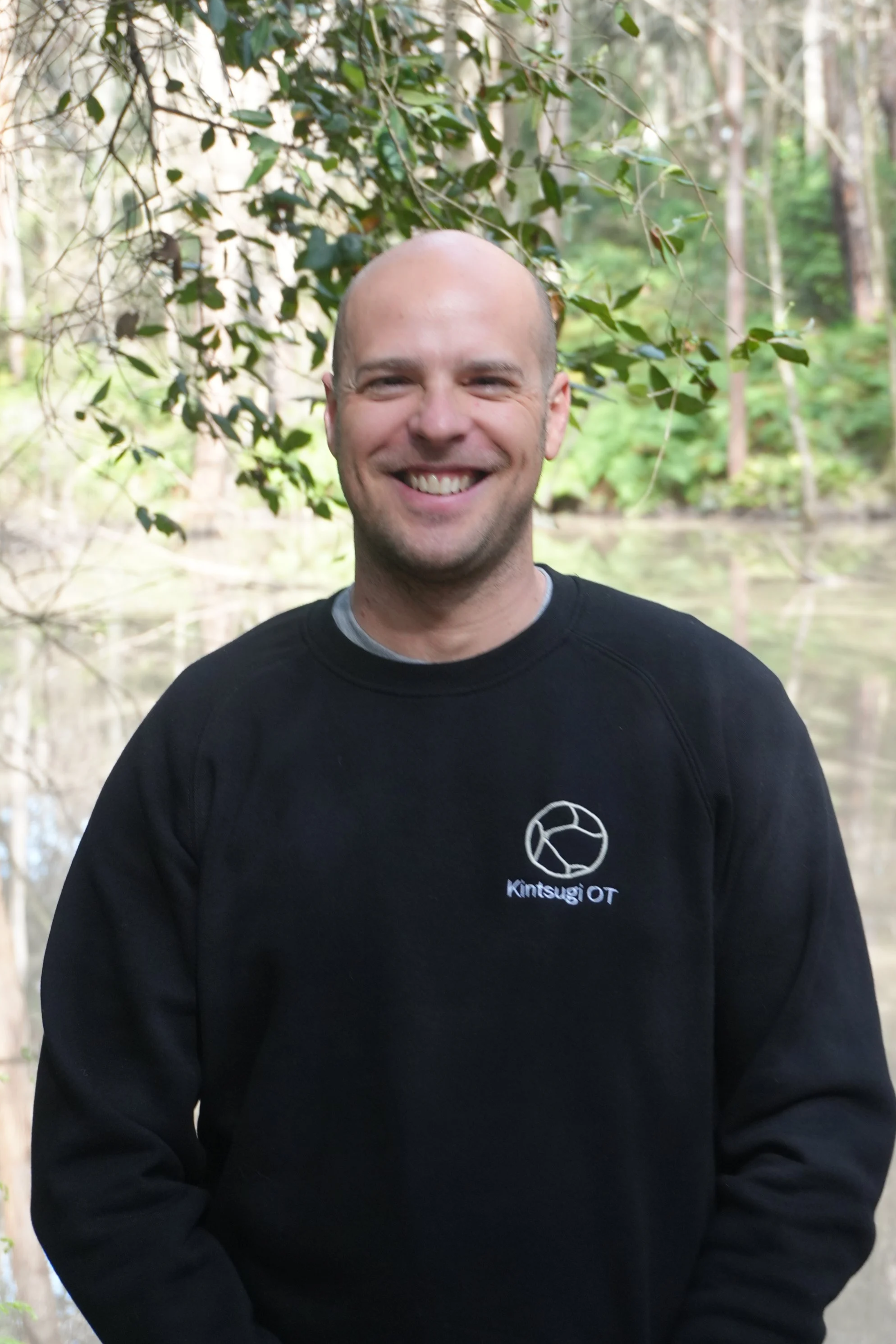 A smiling man standing outdoors near a body of water, surrounded by trees and greenery, wearing a black sweatshirt with a logo that reads "Kintsugi OT."