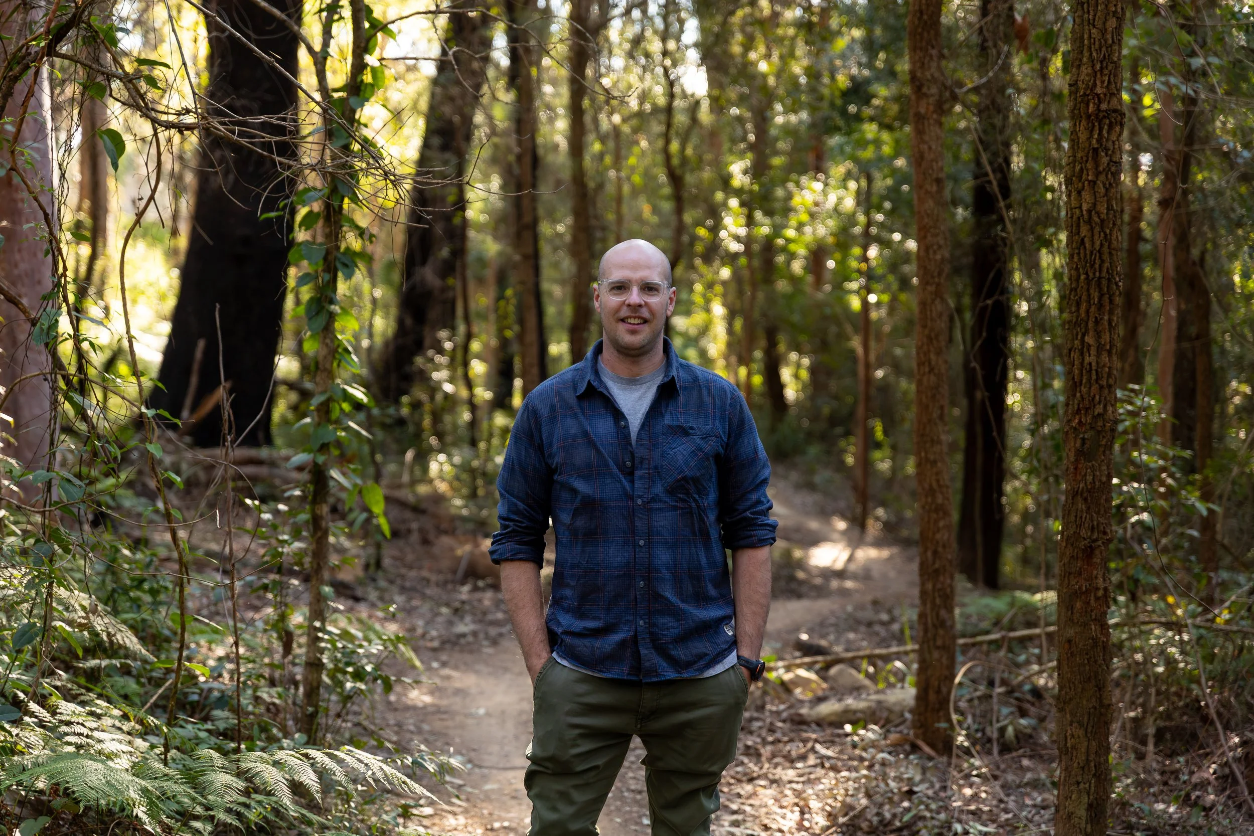 A man stands on a dirt trail in a dense forest, wearing glasses, a blue plaid shirt, and green cargo pants, smiling at the camera.