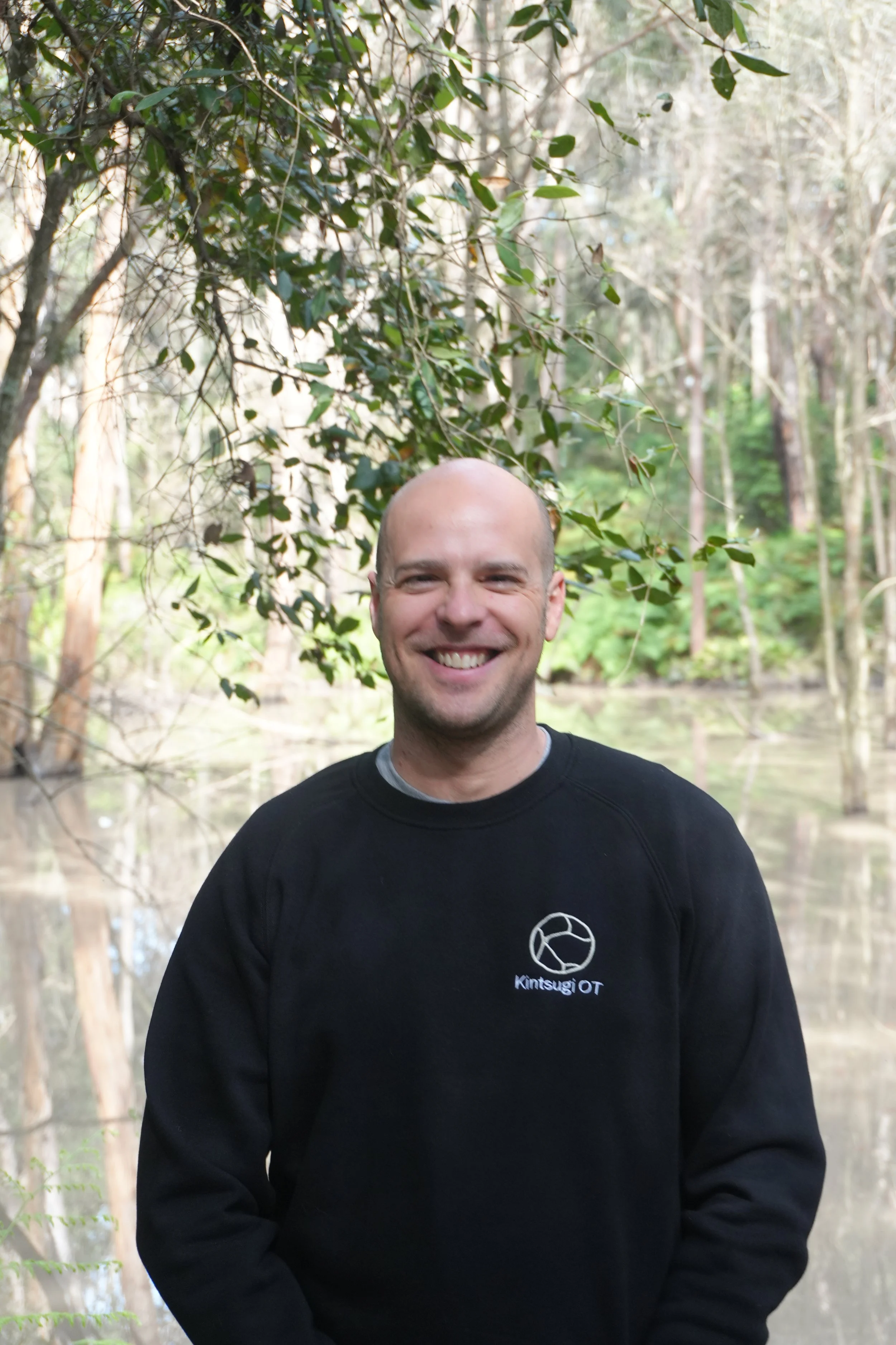A smiling man with a bald head wearing a black sweatshirt with a logo on it, standing outdoors in a wooded area near a body of water.