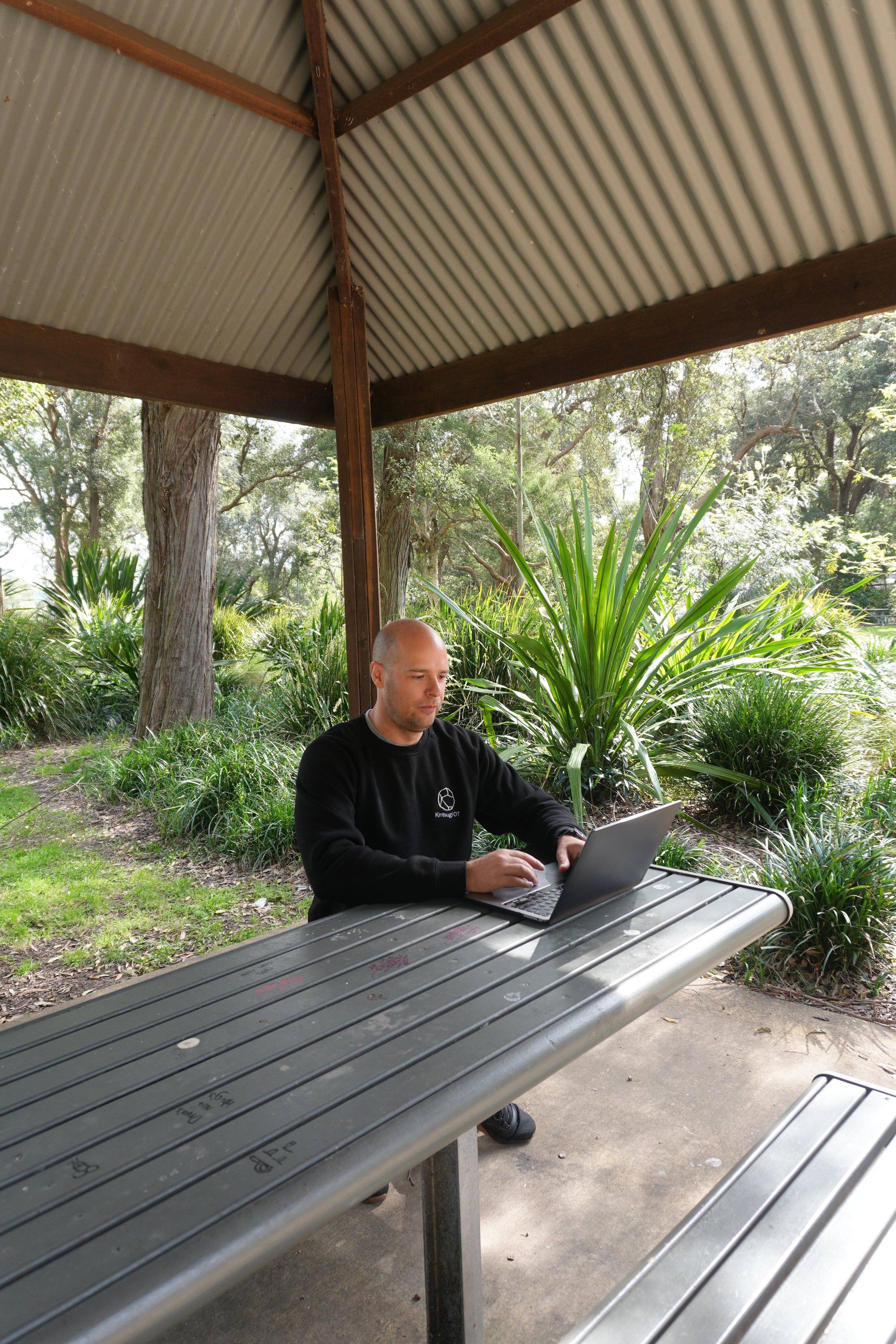 A man working on a laptop at an outdoor picnic table under a metal shelter, surrounded by trees and green foliage.