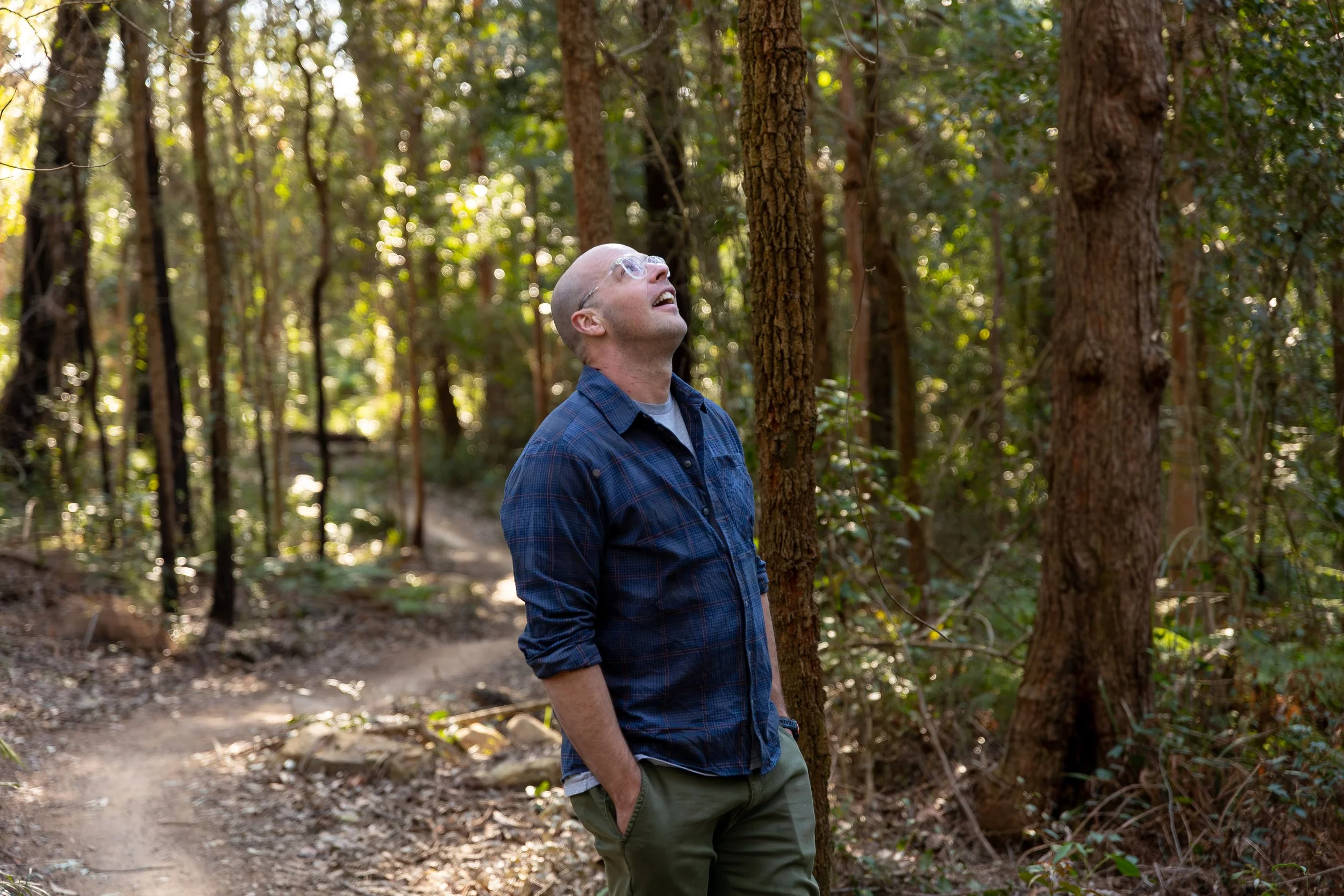 A bald man wearing glasses, a blue checked shirt, and green pants is standing outdoors in a wooded area, looking up at the trees with a joyful expression.