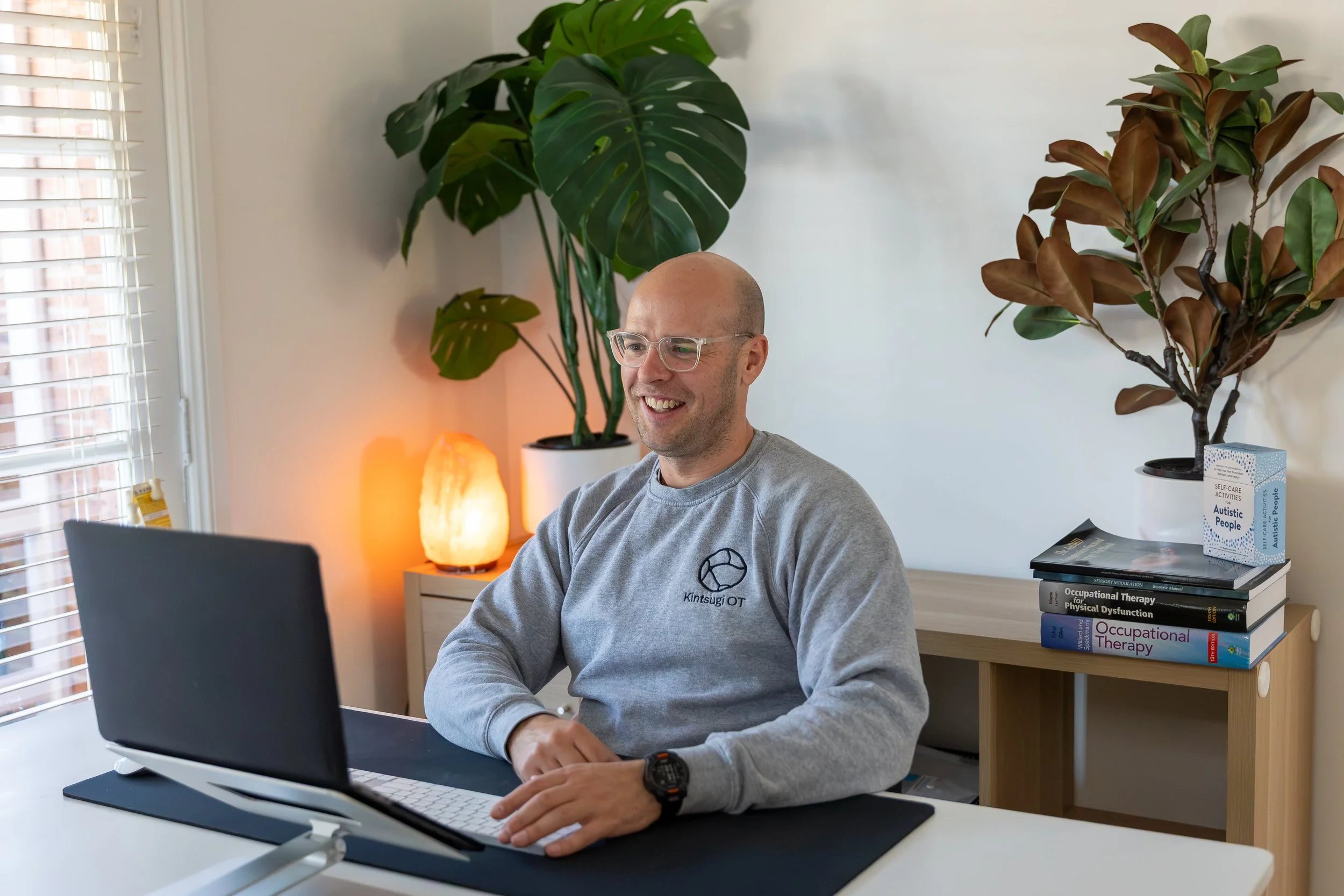 A smiling man with glasses and a salt-and-pepper beard, wearing a gray shirt with a logo, working on a laptop at a white desk in a bright room with a window, houseplants, salt lamp, and books about occupational therapy and autism on the shelves behind him.