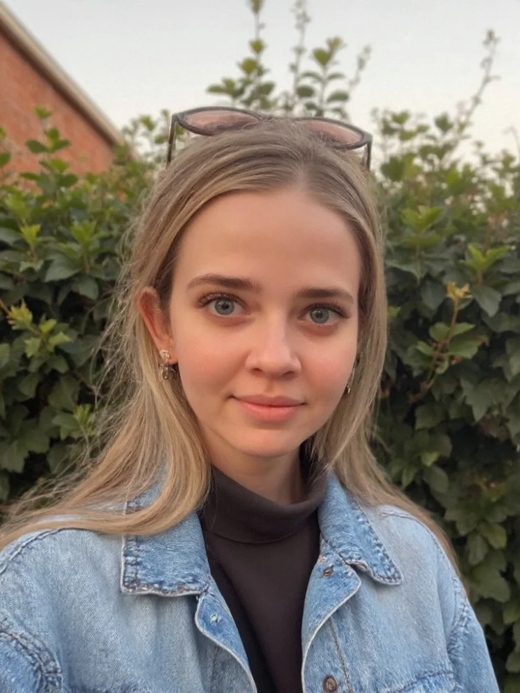 Young woman with blonde hair and blue eyes, wearing sunglasses on her head, a black turtleneck, and a denim jacket, standing outdoors in front of green bushes and a building.