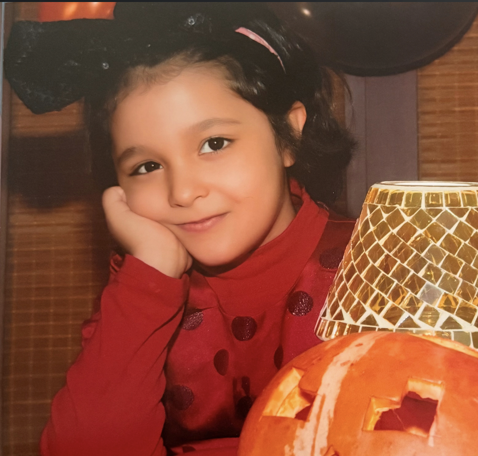 A young girl with dark hair and light brown skin, smiling with her hand resting on her cheek, wearing a red top with polka dots, sitting at a table with a carved pumpkin and a decorative lamp, in a cozy indoor setting.