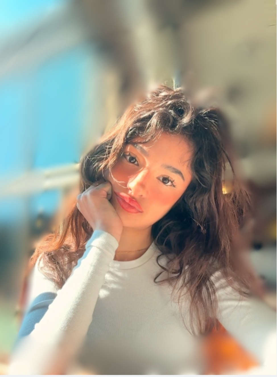 A woman with wavy brown hair resting her chin on her hand, looking at the camera, indoors with warm lighting.