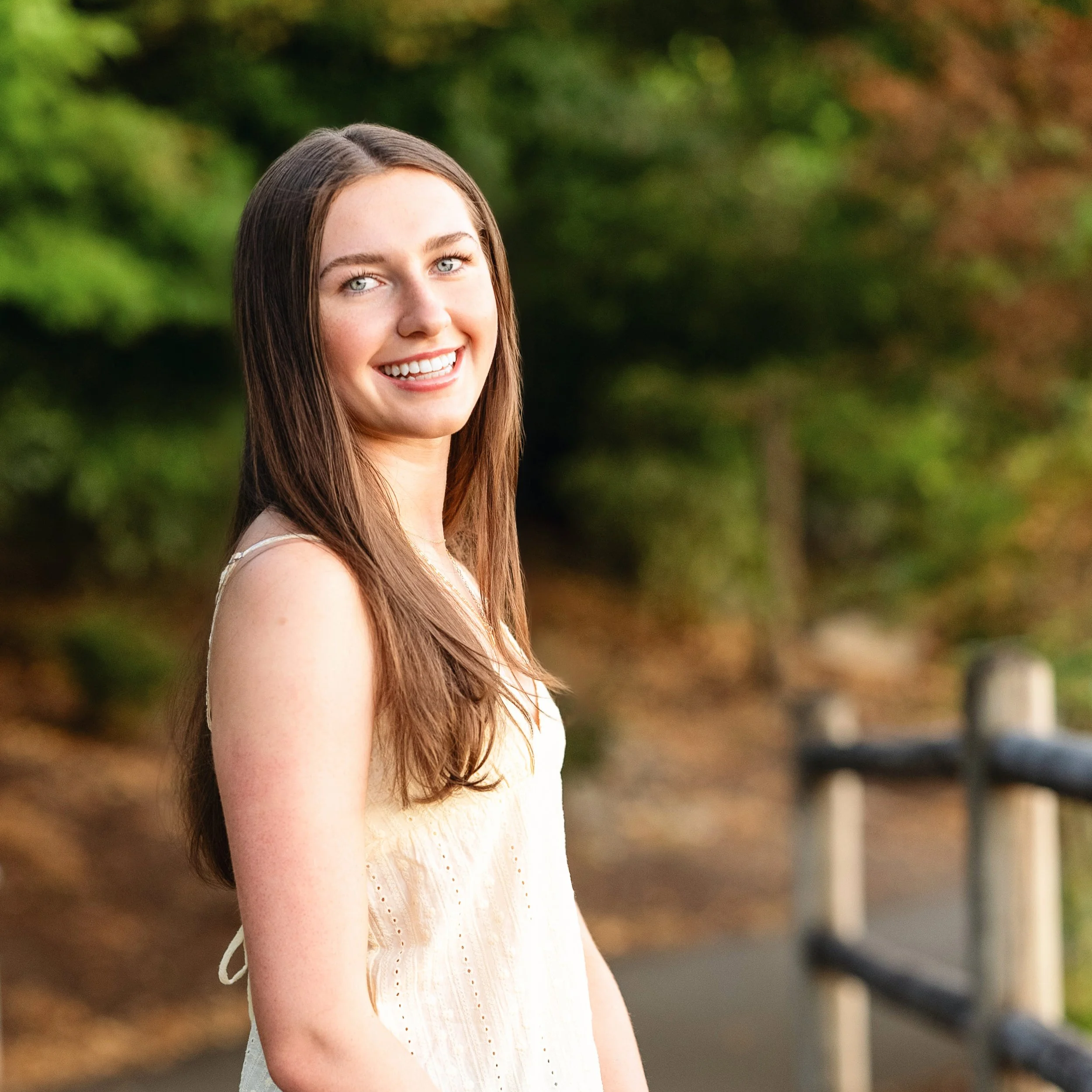 A young woman with long brown hair and blue eyes smiling outdoors near a forested area with a wooden fence.