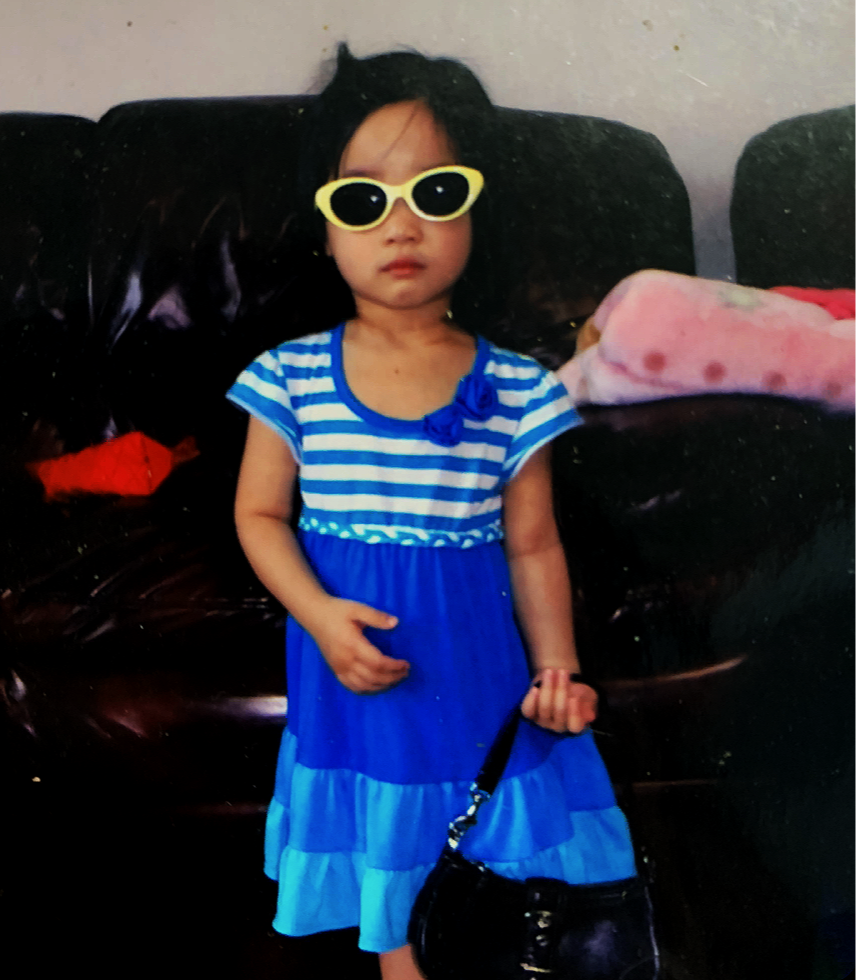 Young girl with dark hair wearing yellow sunglasses and a blue striped dress, holding a small black purse, standing in front of a black leather couch with a pink blanket on it.