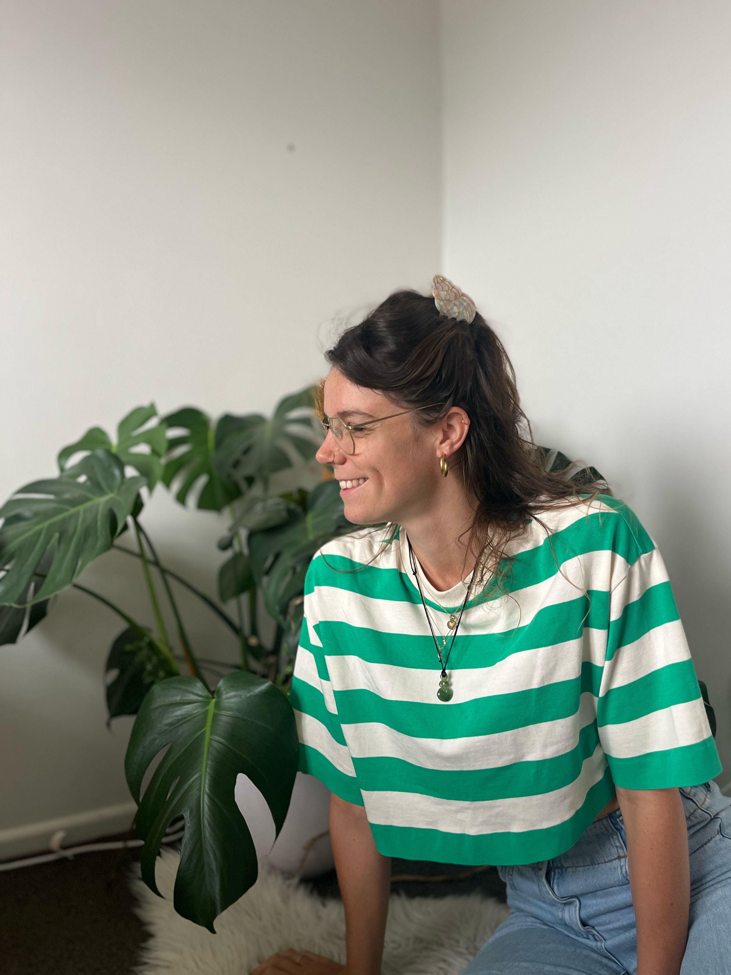 A woman with dark wavy hair, glasses, and hoop earrings, smiling while sitting on a fluffy white rug next to a large green monstera plant. She is wearing a green and white striped shirt and jewelry, including necklaces and a hair clip.