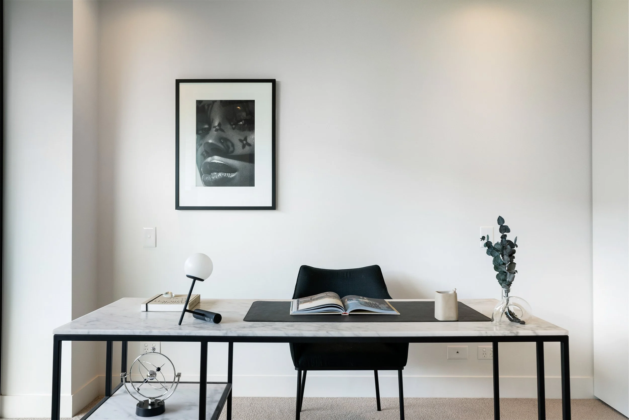 Modern minimalist office with a white marble desk, black chair, and wall art featuring a black-and-white photo of a woman with face markings.