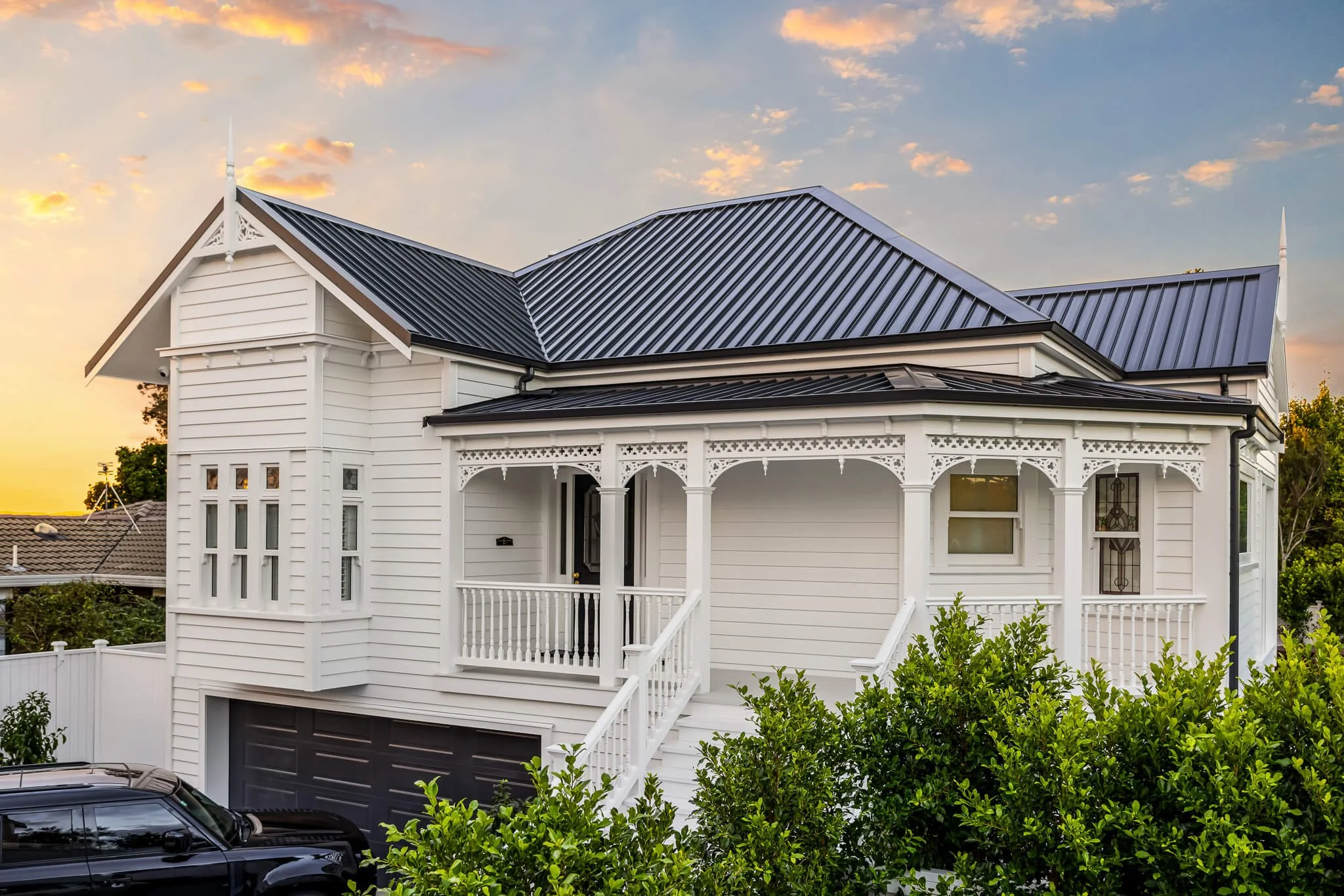 A white Victorian-style house with decorative trim, multiple windows, and a black garage door, surrounded by greenery, at sunset.