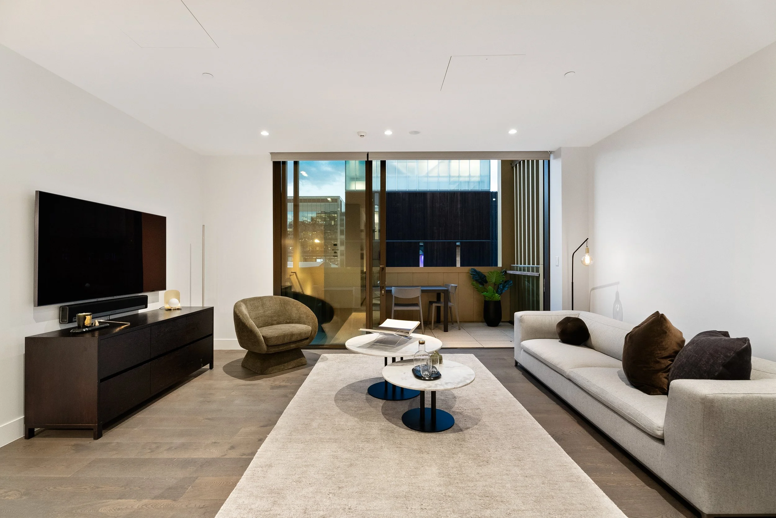 Modern living room with beige sofa, round marble coffee tables, TV on a black console, and a balcony with outdoor seating and potted plant.