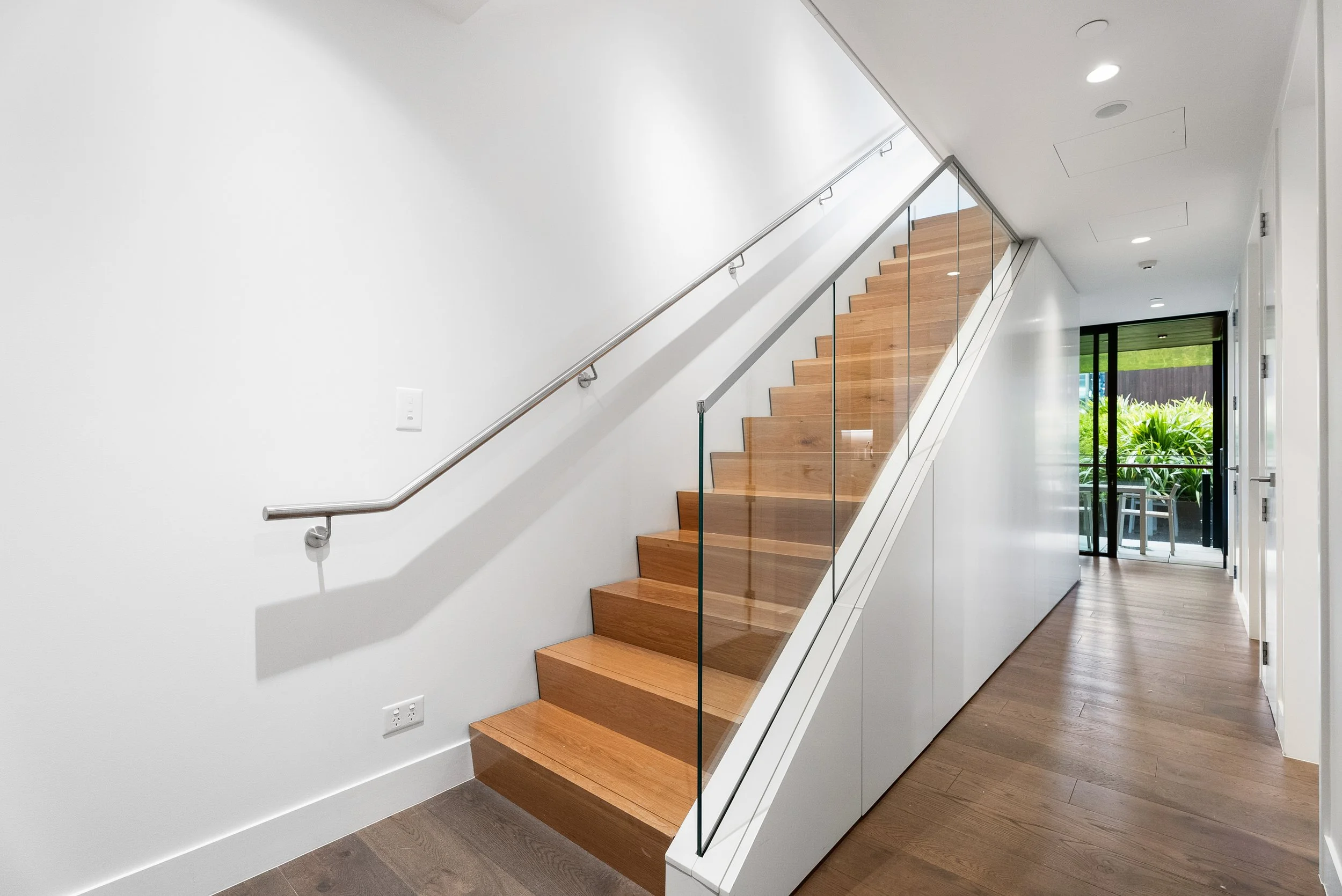 Interior of a modern house with wooden stairs, a glass railing, white walls, and a door leading outside to a patio with green plants.