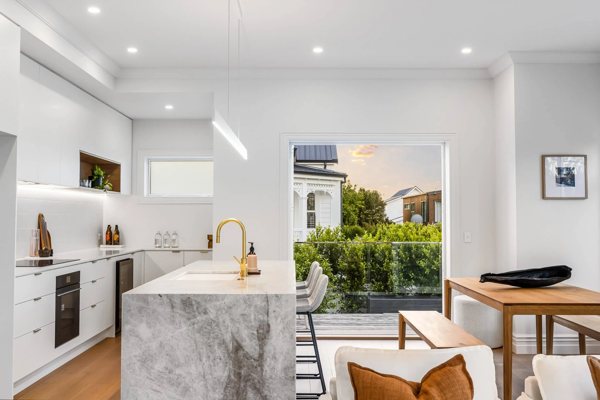 Modern kitchen with white cabinets, marble island with gold faucet, open view of balcony and greenery outside, and living area with beige furniture and throw pillows.