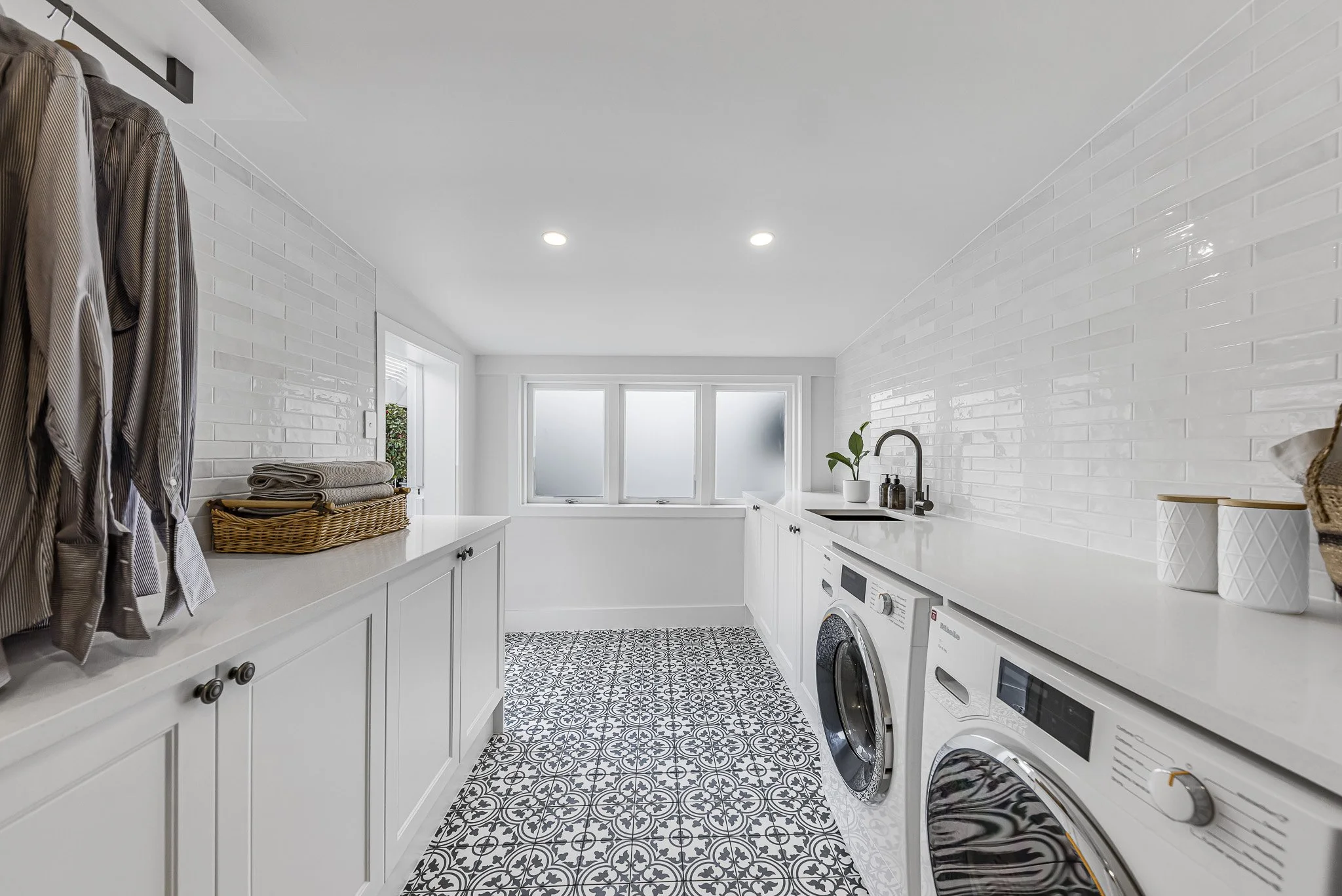 A bright laundry room with white cabinetry, patterned black and white tile flooring, and a row of three frosted windows. Contains a washing machine and dryer, a sink with a black faucet, plants, and decorative items.