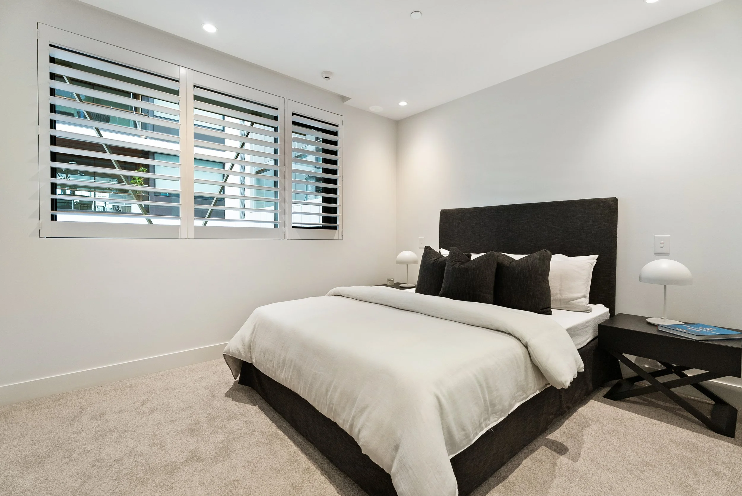 A modern bedroom with a bed, black headboard, black pillows, white sheets, and beige carpet. There are white nightstands with white lamps on each side and a window with horizontal blinds.