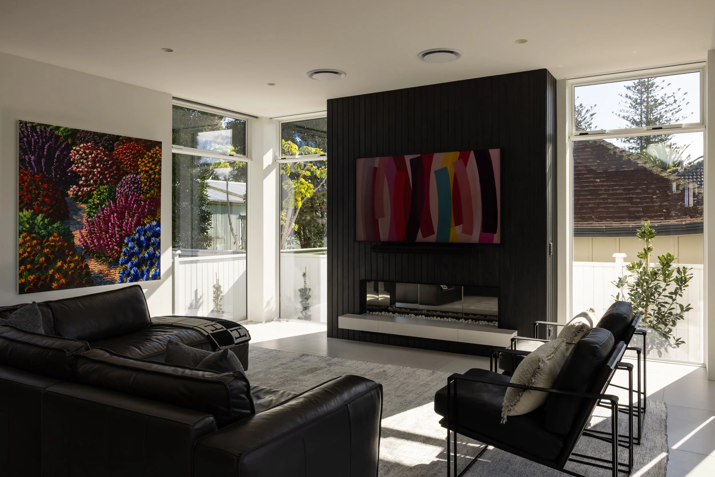 Modern living room with black leather sofas, colorful abstract art on the walls, and large windows letting in sunlight.