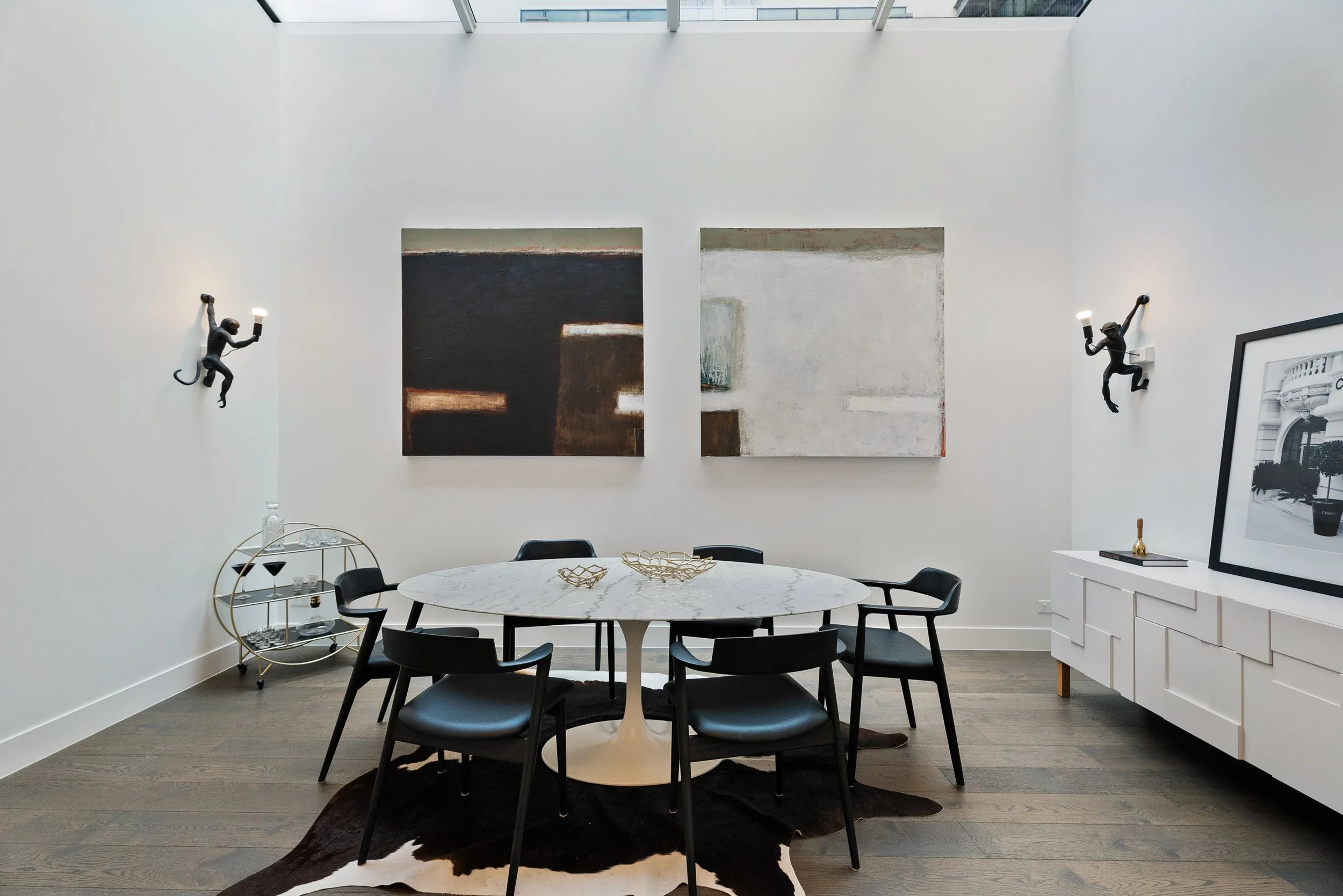 Modern dining room with white walls and a skylight ceiling. Features a round marble table with five black chairs, abstract artwork on the wall, and decorative monkey-shaped wall lamps holding lights.
