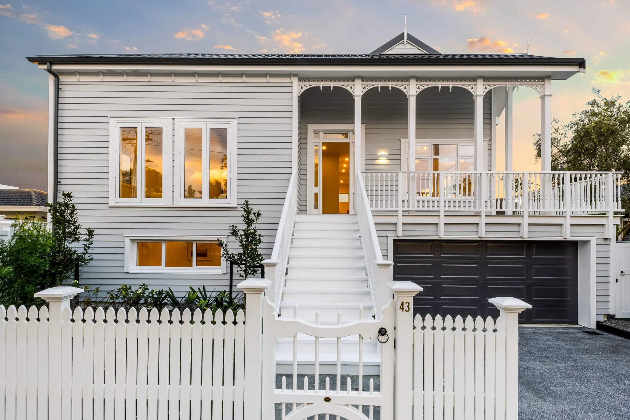 White two-story house with black garage door, front staircase, and decorative porch, surrounded by a picket fence, at sunset.