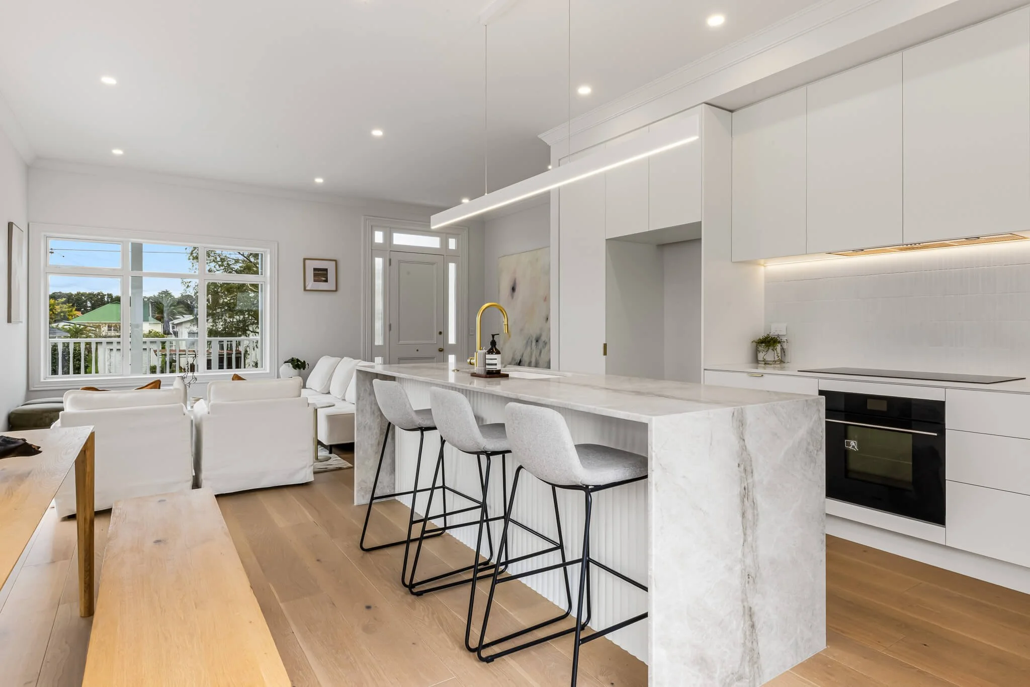 Modern kitchen with white cabinets, marble island, four gray bar stools, and a view of a living area with white sofas and large window