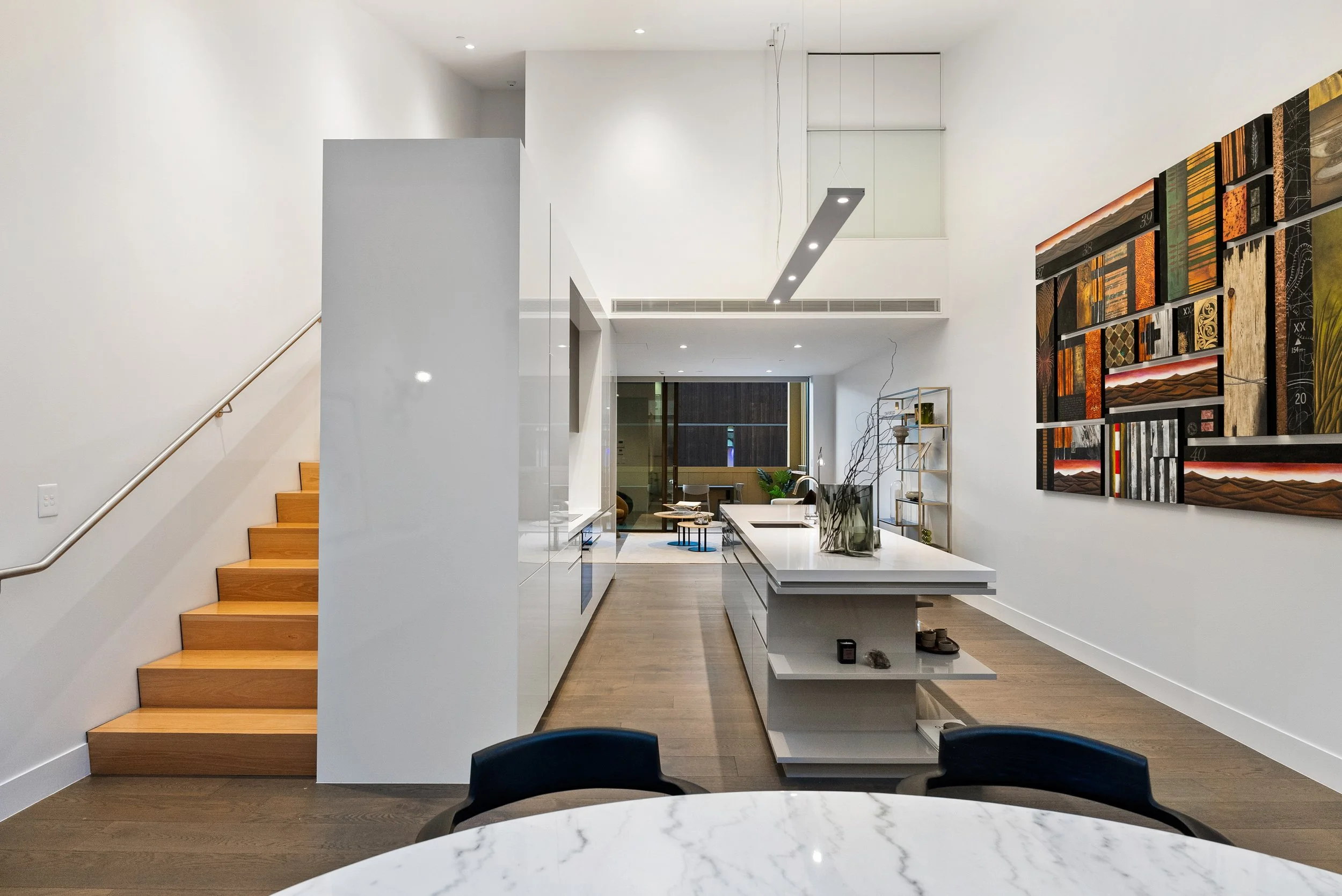 Modern, open-concept kitchen with white cabinetry, a large island, and a wooden staircase on the left. The kitchen has a decor piece with abstract wall art and a nearby sitting area with a window in the background.