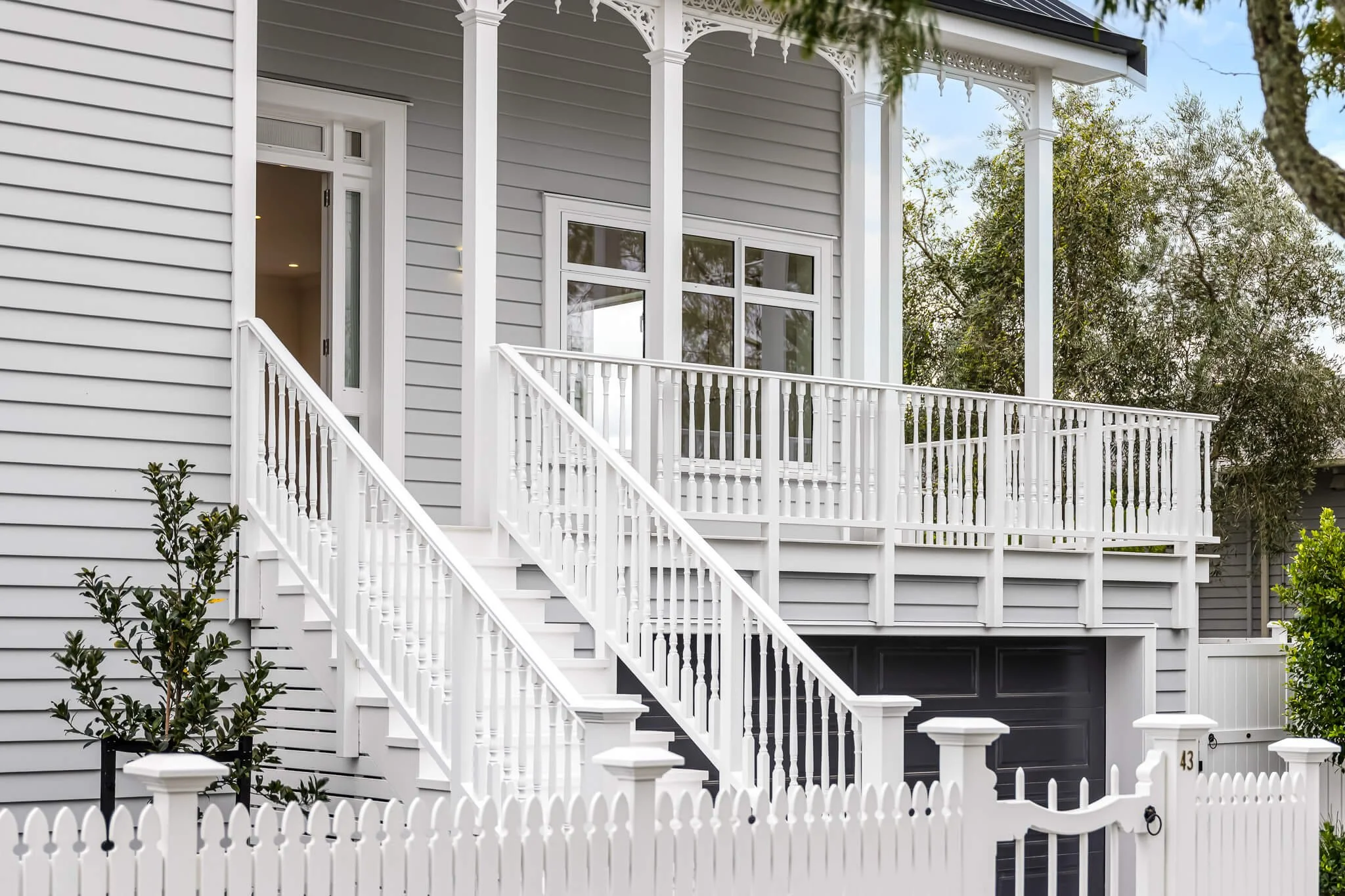 Front view of a modern, gray house with white porch railings, stairs, and fencing, featuring a large window, a partially open door, and decorative trim, with trees and greenery in the background.
