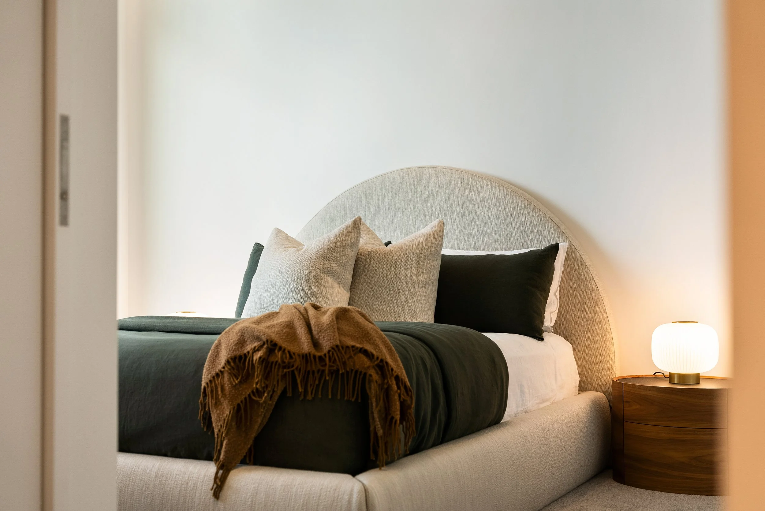 A neatly made bed with dark green, white, and black pillows, a beige headboard, a brown throw blanket, and a wooden nightstand with a white lamp on it, in a cozy bedroom.