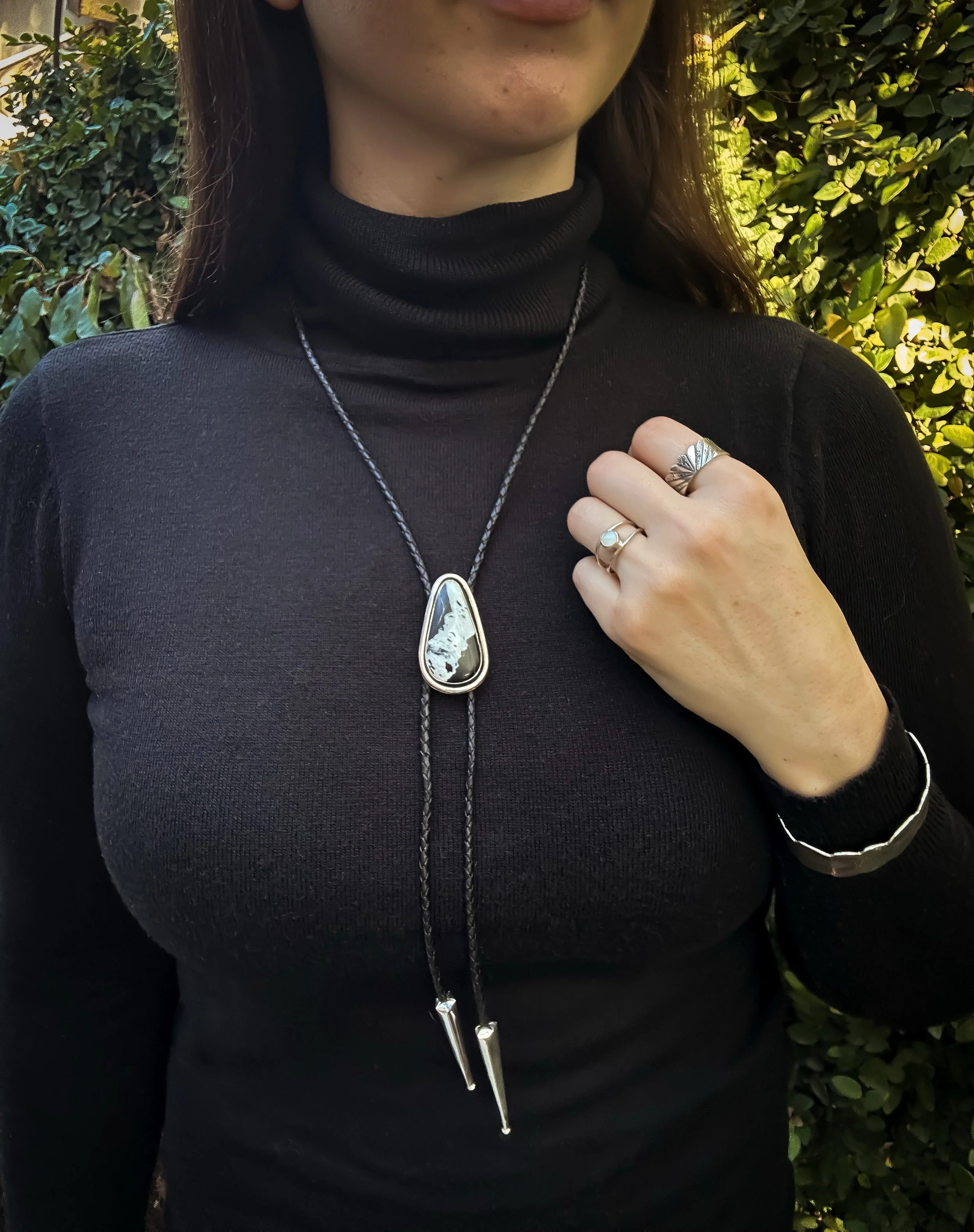 Woman wearing a black turtleneck, with silver and gemstone jewelry, standing outdoors against a leafy background.