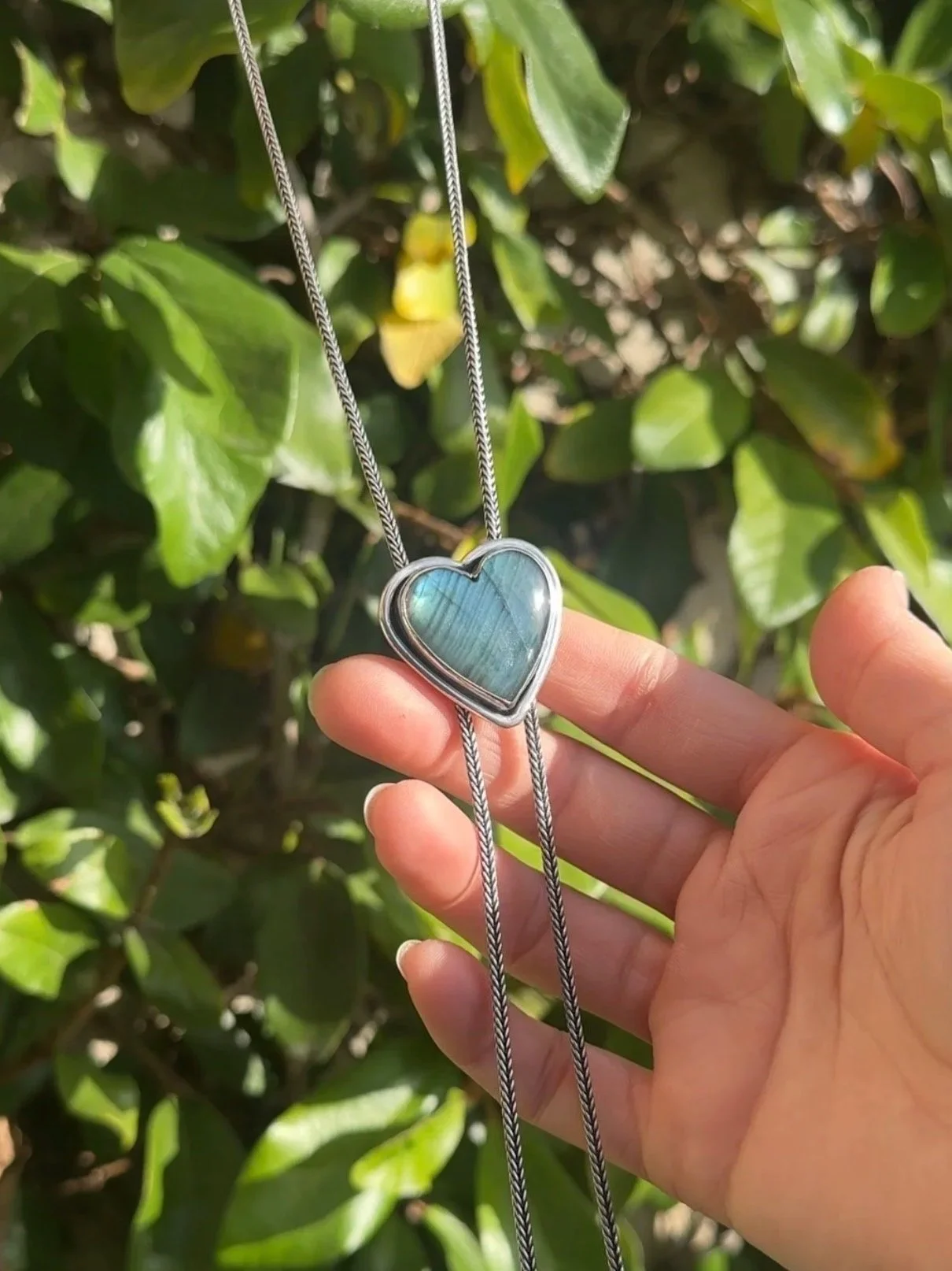 Person holding a necklace with a heart-shaped pendant featuring a blue-green stone, against green leafy background.