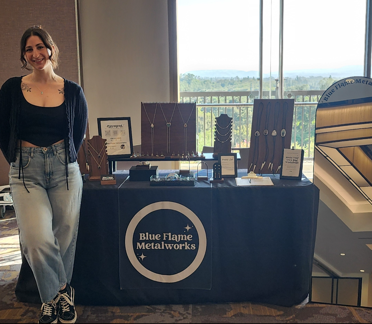 A woman standing next to a jewelry display table with necklaces, bolo ties, rings, and bracelets, titled Blue Flame Metalworks, inside a room with large windows showing a scenic view.