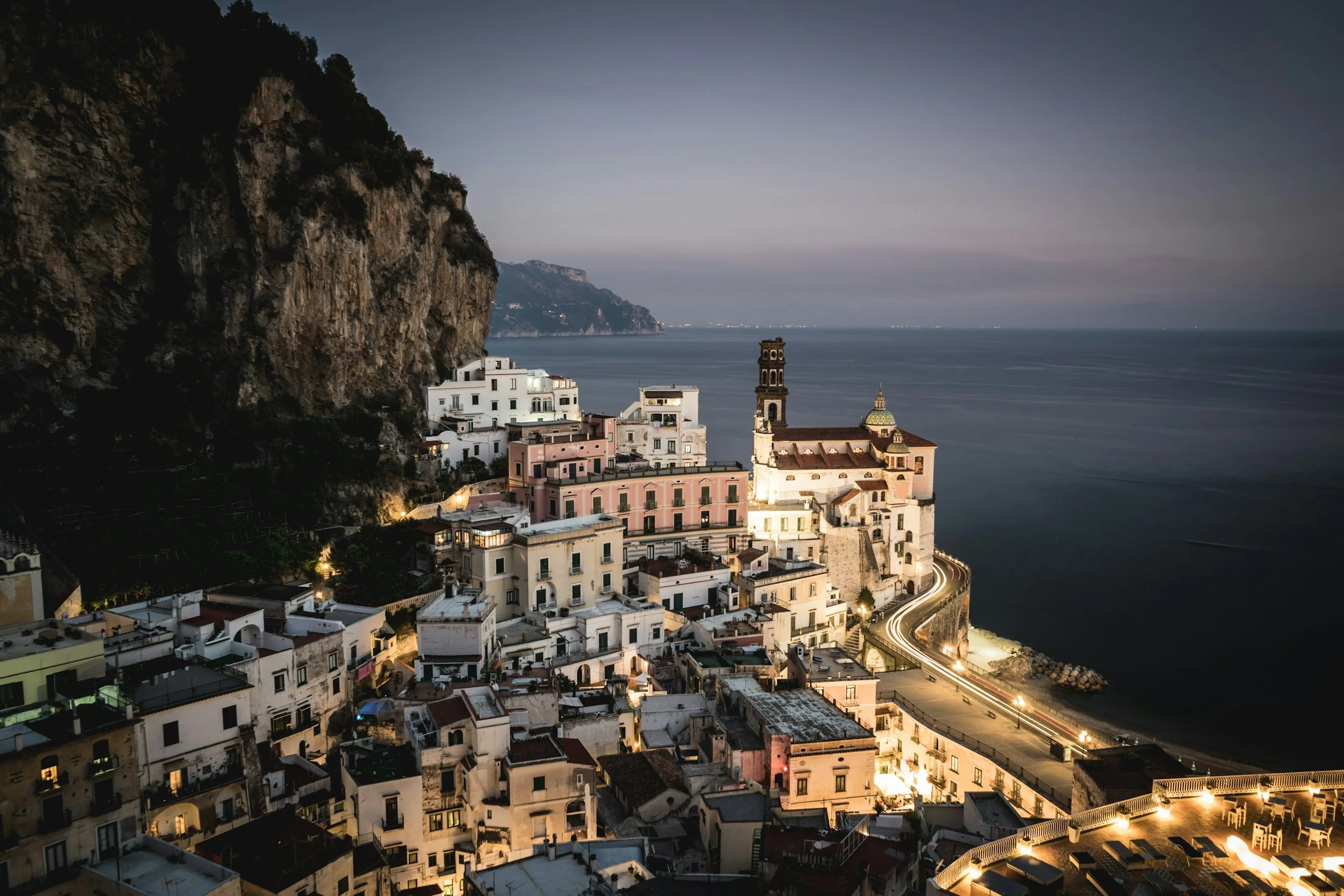 A coastal village with white buildings on a hillside overlooking the sea at dusk, with a church tower and a winding road illuminated by streetlights.